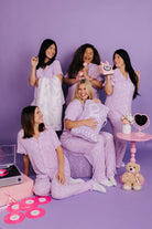 Five women in matching lavender pajamas posing against a purple background with various props.