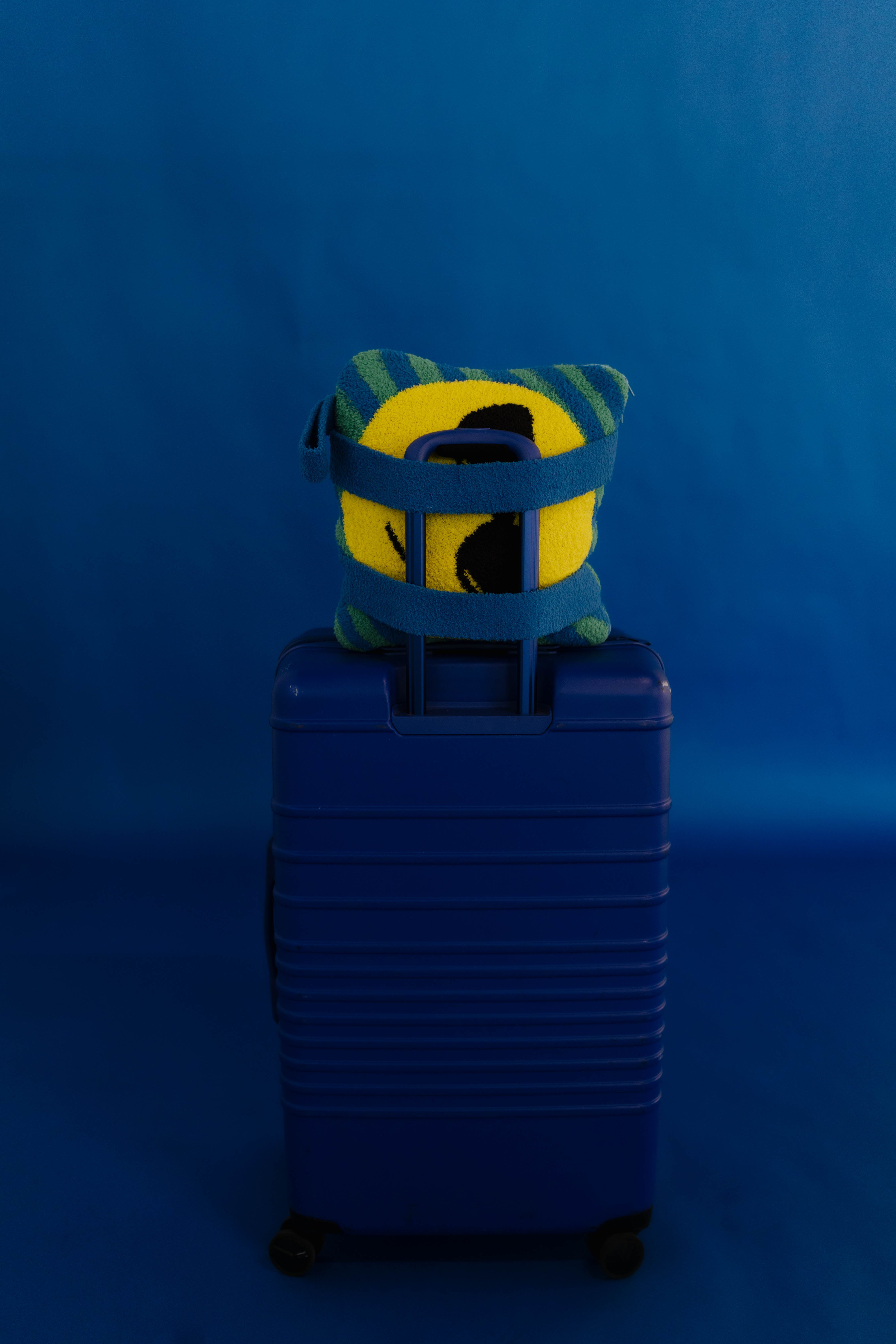 A blue hard-shell rolling suitcase standing upright against a blue studio background, with a yellow and green striped plush quillow tucked into the top handle area.