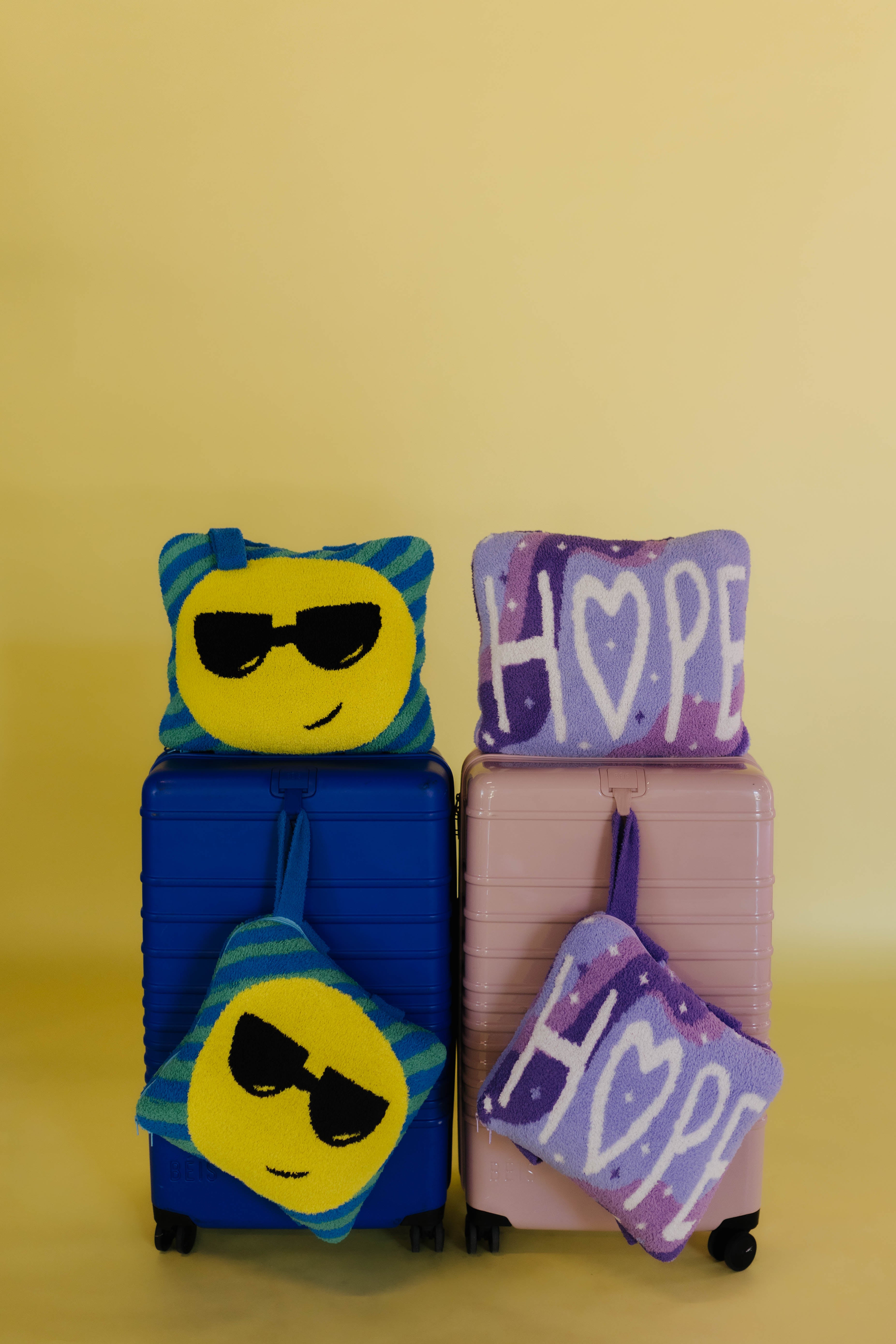 Two hard-shell suitcases, one blue and one pink, each with quillows on them. The blue suitcase has a yellow smiley face with black sunglasses quillow on top and a purple 'HOPE' pouch below.