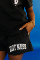 Person wearing a black outfit with 'HOT MESS' shorts against a blue background
