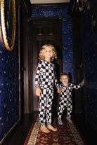 Two children standing in a dark hallway wearing matching black-and-white checkered pajamas. The room has dark blue patterned wallpaper, a dark wooden door behind them, and a red patterned runner rug.