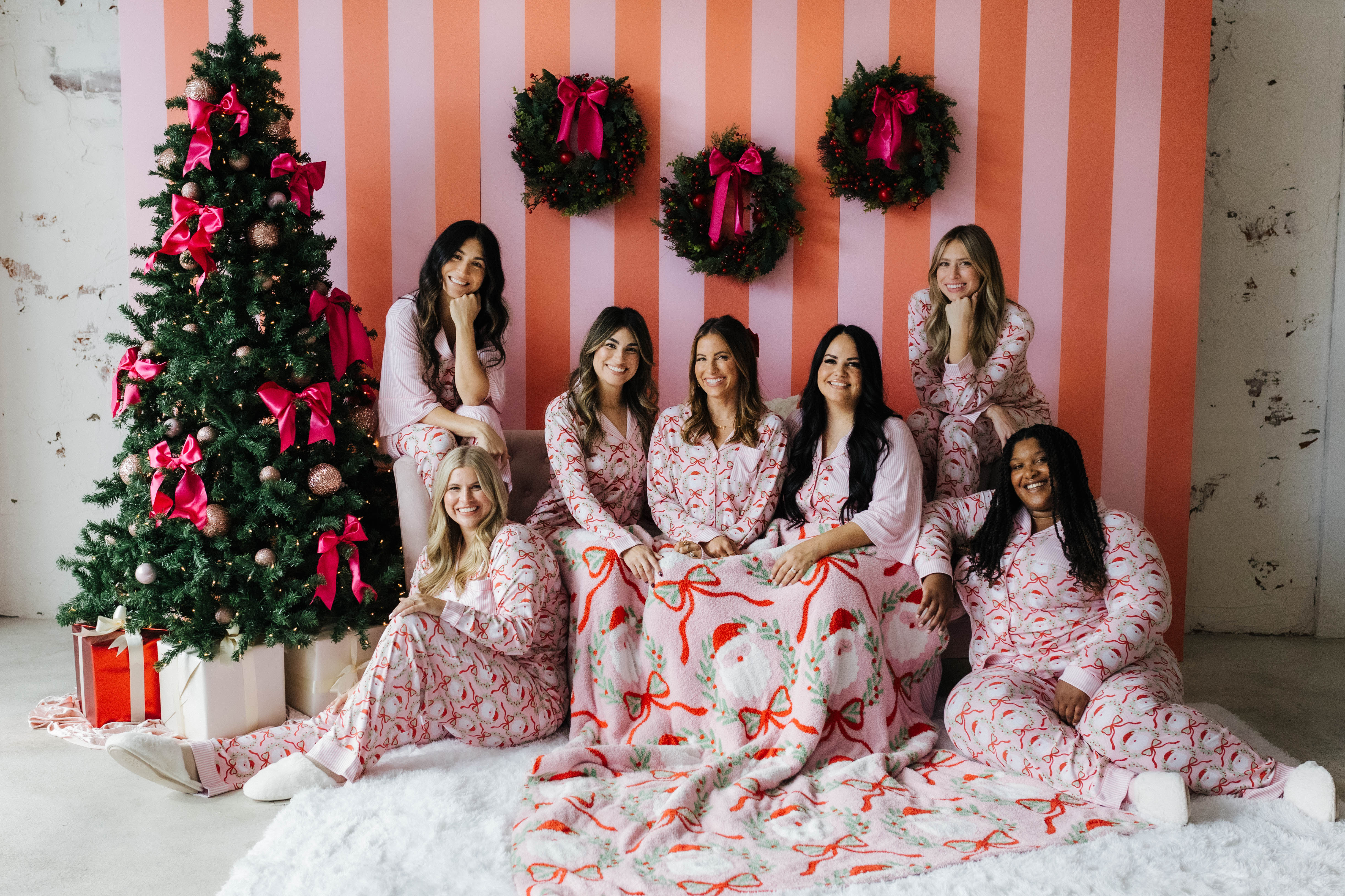 Group of women in matching pajamas posing in front of a Christmas tree and striped wall.