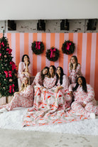 Group of women in matching pajamas sitting on a white blanket with a Christmas tree and striped wall in the background.