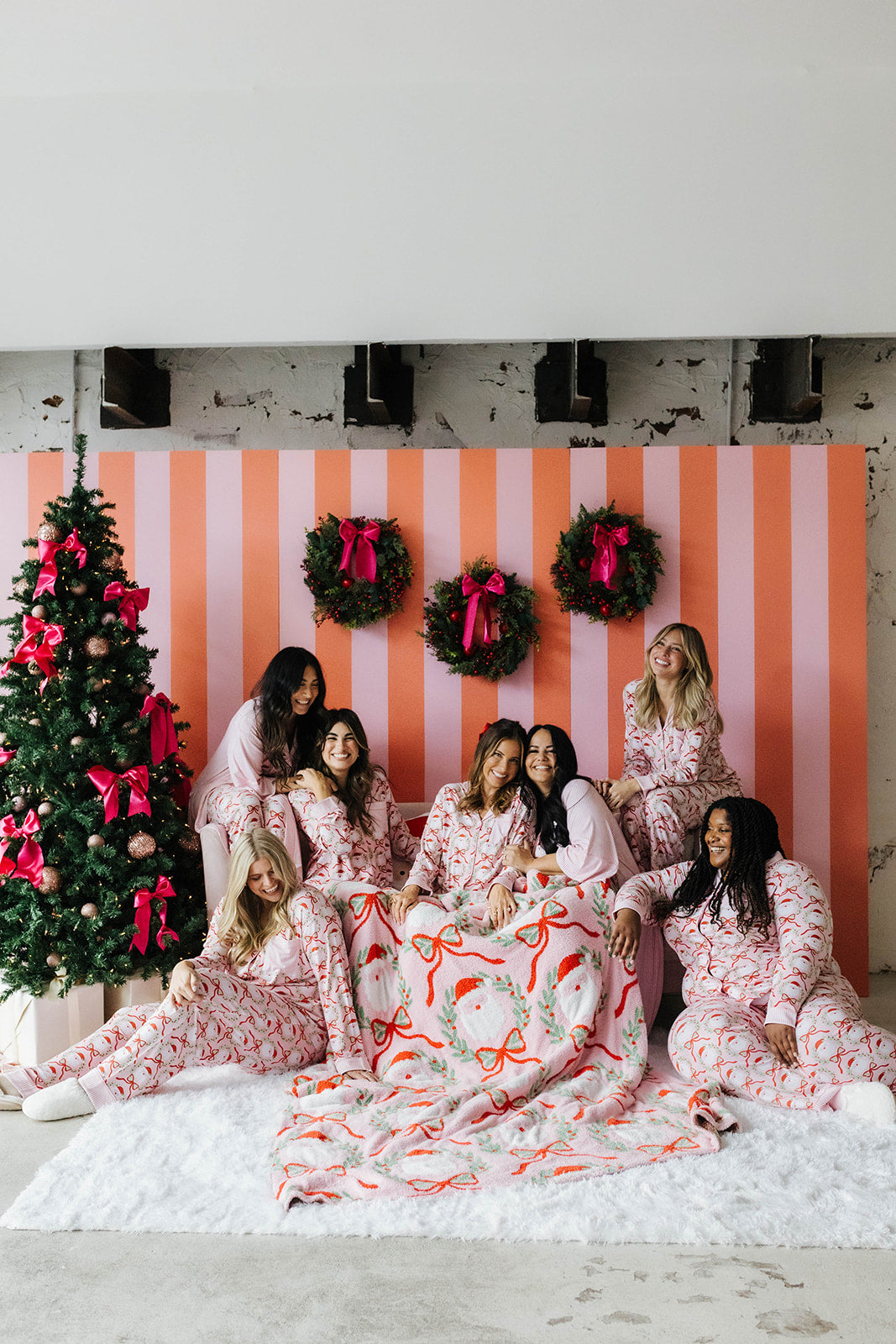 Group of women in pajamas sitting on a white rug in front of a Christmas tree and striped wall.