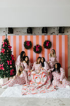 Group of women in pajamas sitting on a white rug in front of a Christmas tree and striped wall.