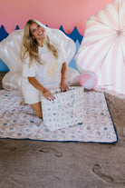 Woman setting up a beach-patterned blanket on sand with colorful inflatables in the background.