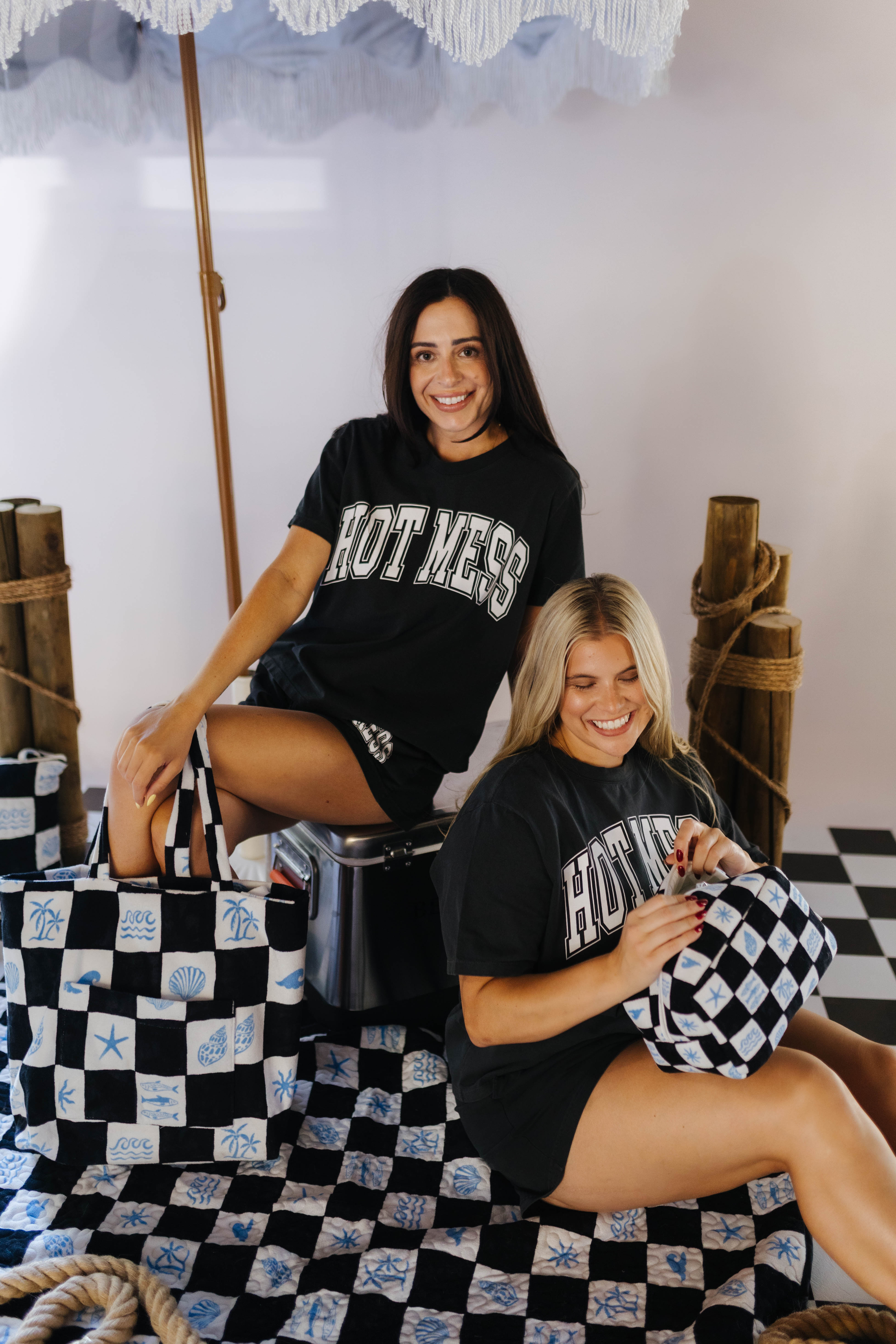 Two women sitting on a checkered floor with black and white bags, wearing matching 'HOTMESS' t-shirts.