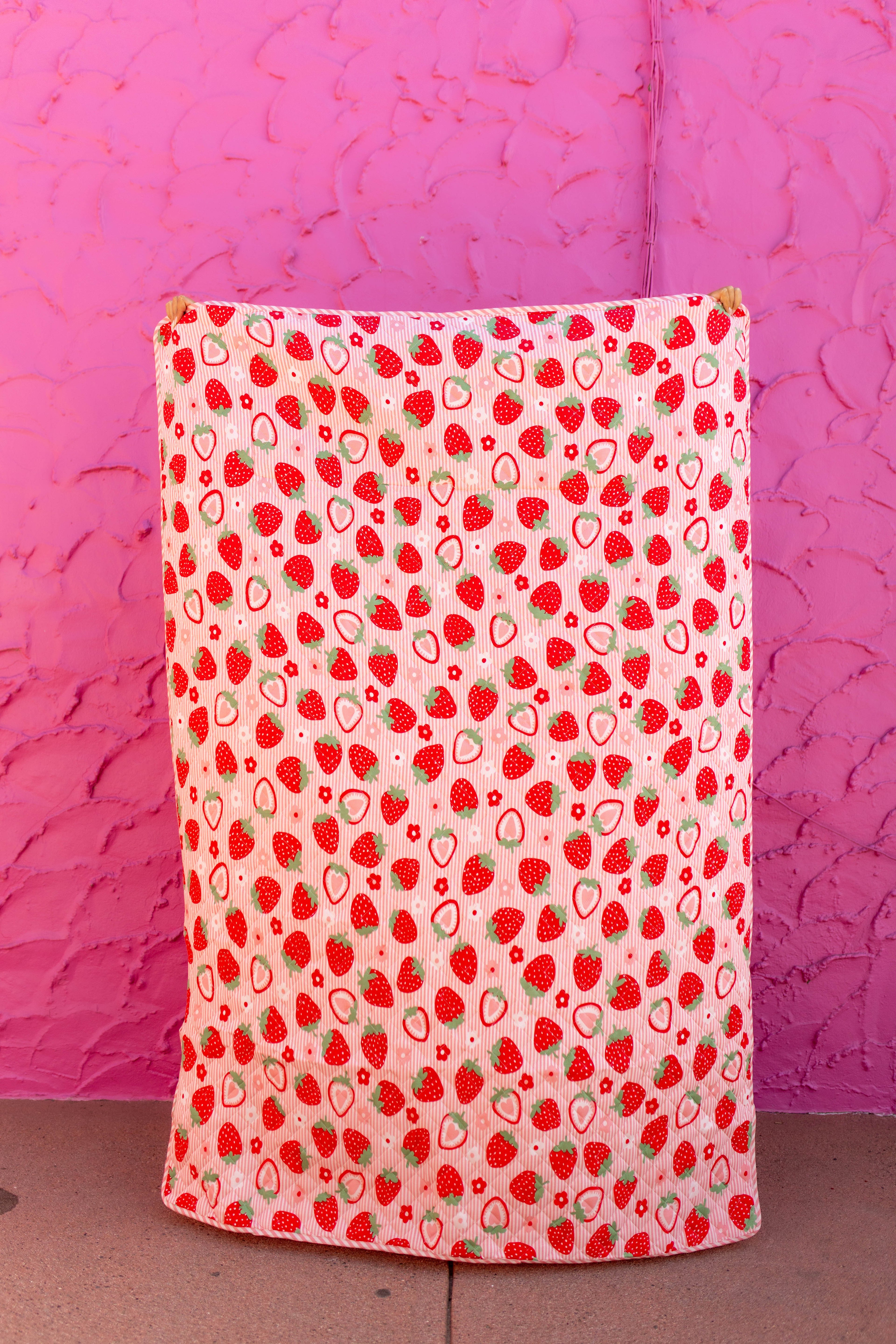 the front of the outdoor quillow with strawberry and subtle pink stripe background held up against a hot pink background
