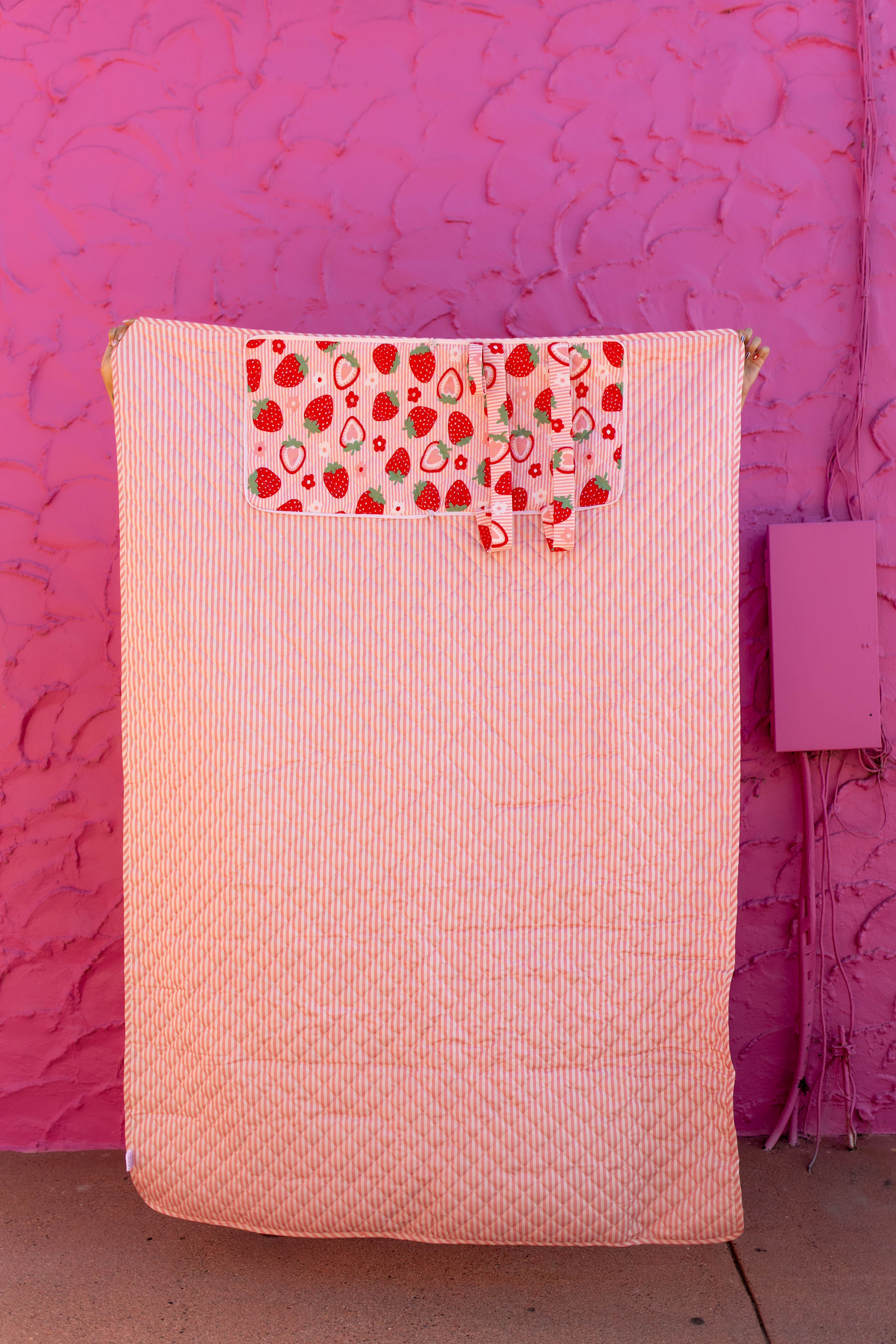 A quilted striped-pink outdoor blanket with a decorative pocket featuring a strawberry and fruit print, set against a textured hot pink wall.