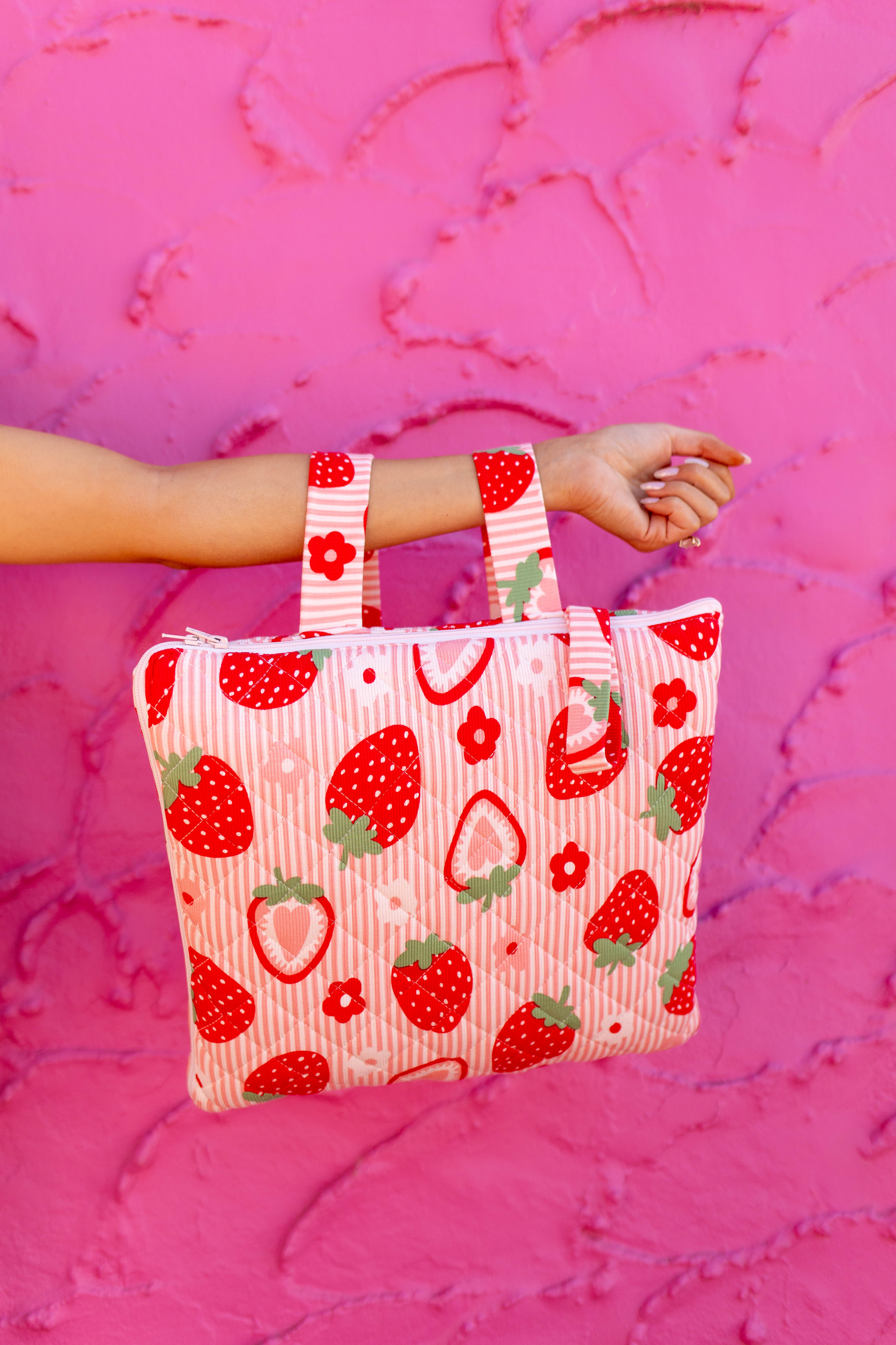 A striped fabric strawberry and flower print quillow held by a person's arm against a textured hot pink background.