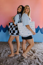 Two women holding patterned tote bags on a beach with a colorful background