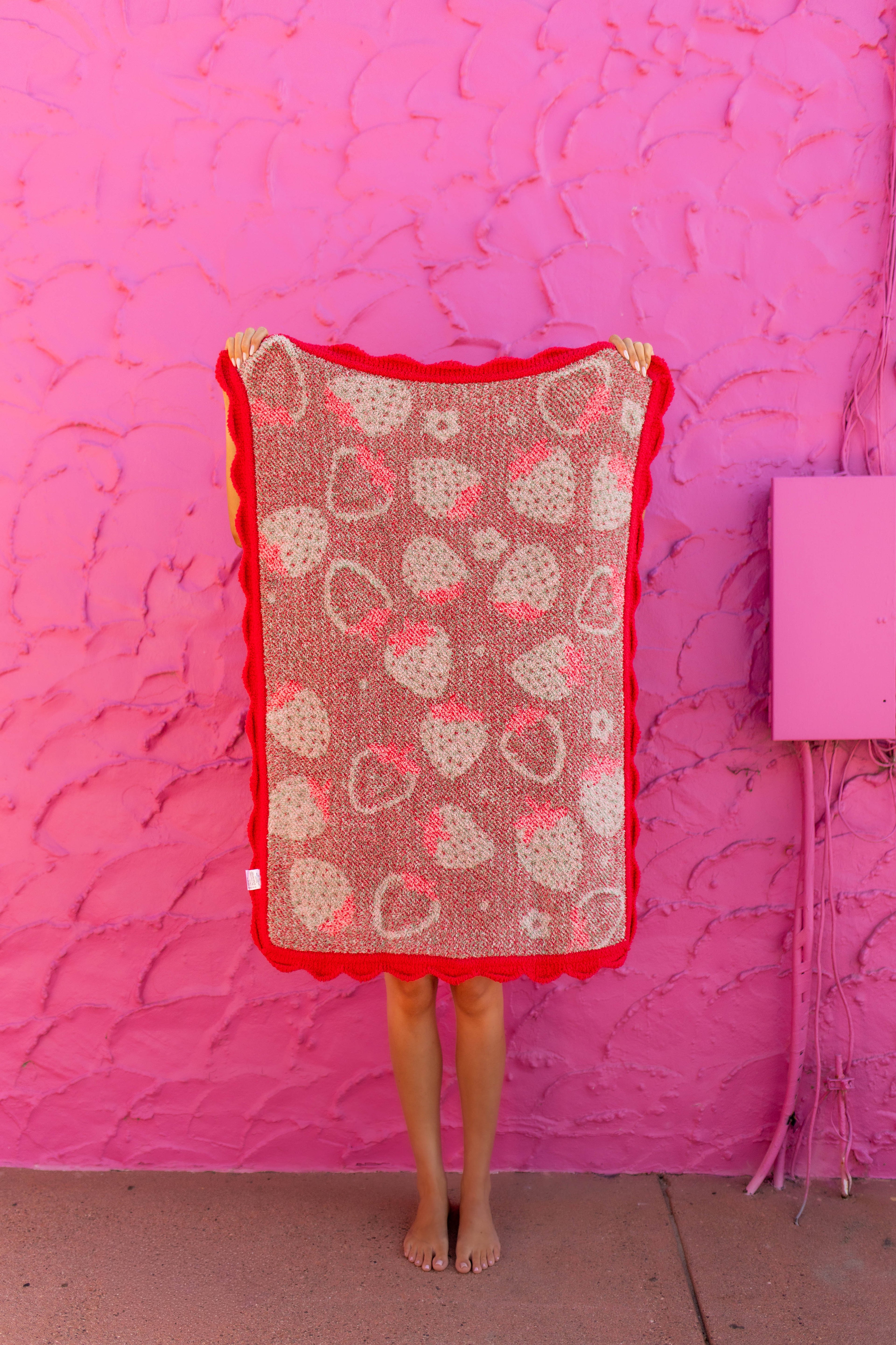 A person standing barefoot in front of a bright pink textured wall, holding up a rectangular crochet blanket with a red border and a pattern of pink and beige hearts.