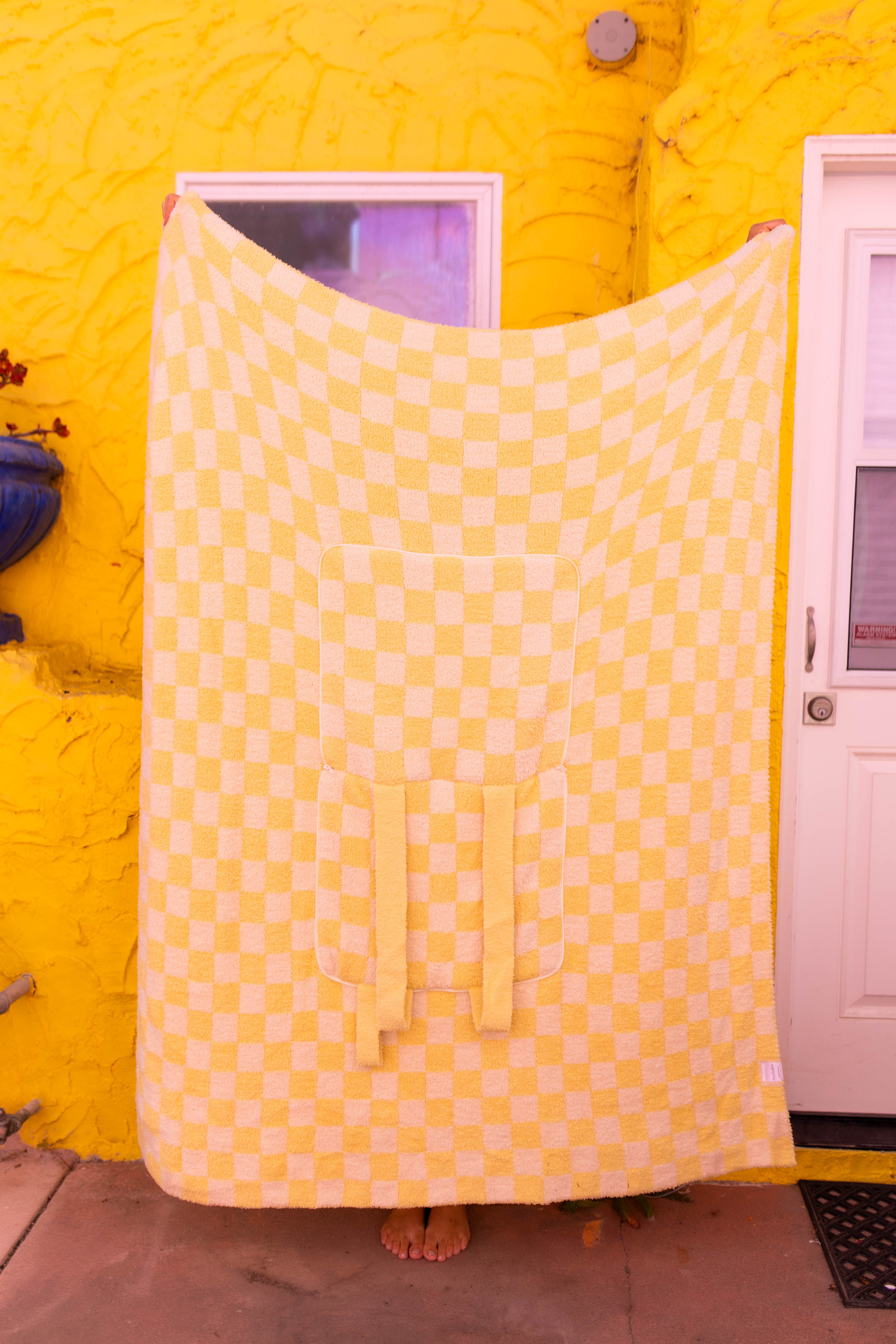 A large checkered yellow quillow is held up by a person standing barefoot in front of a bright yellow stucco wall with a white door.