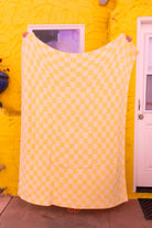 A large yellow checkered quillow is held up by a person in front of a bright yellow wall with a white door visible on the right.