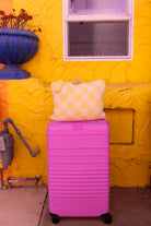 A bright pink hard-shell suitcase with a yellow checkered quillow on top, set against a textured yellow stucco wall with a blue planter and window.