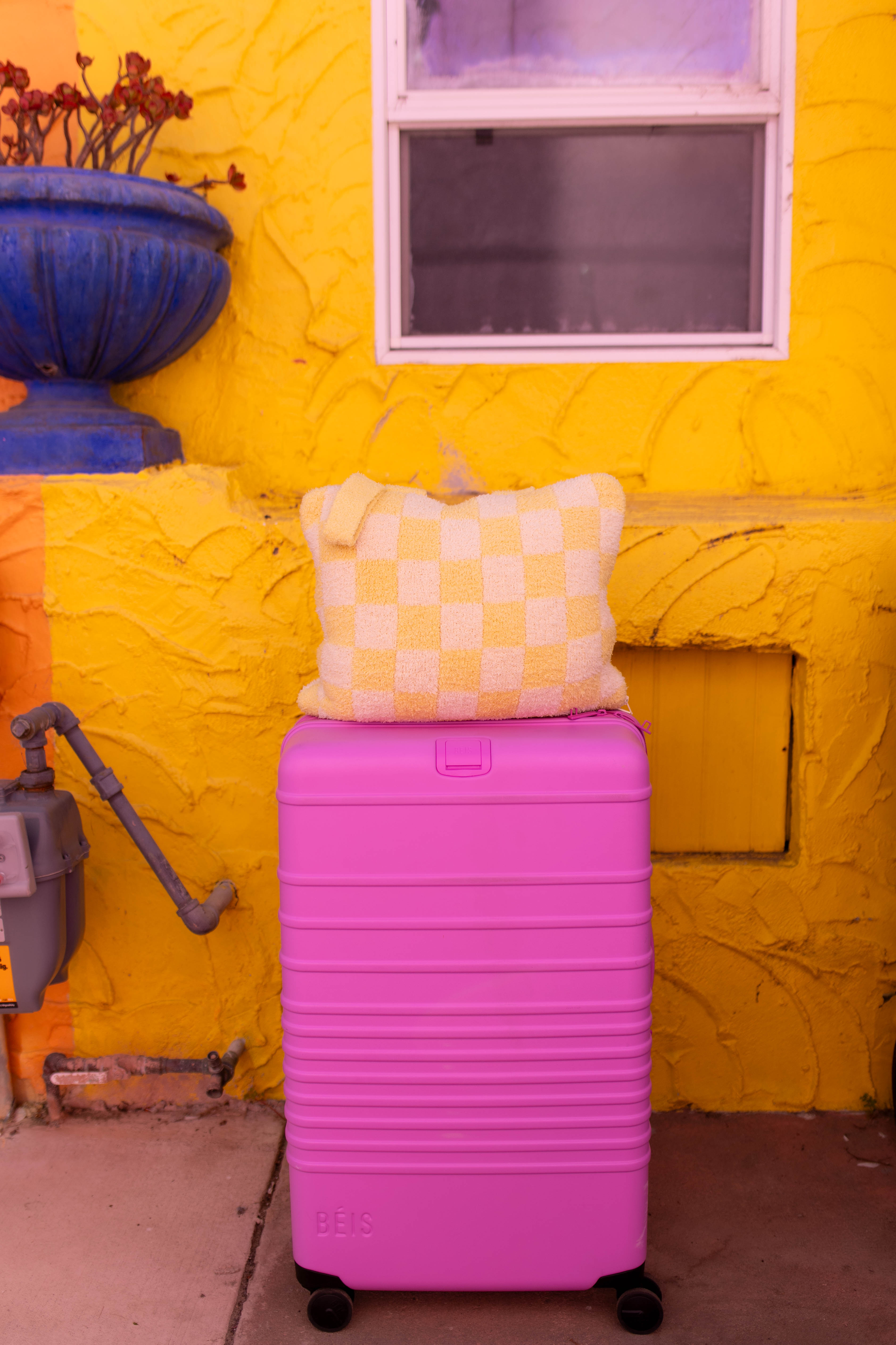 A bright pink hard-shell suitcase with a yellow checkered quillow on top, set against a textured yellow stucco wall with a blue planter and window.