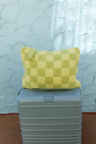 A light green checkered plush quillow with a handle, resting on top of a gray hard-shell suitcase against a textured light blue wall.