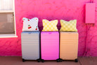 Three hard-shell rolling suitcases in pastel blue, pink, and tan are lined up against a bright pink textured wall, each topped with a quillow.