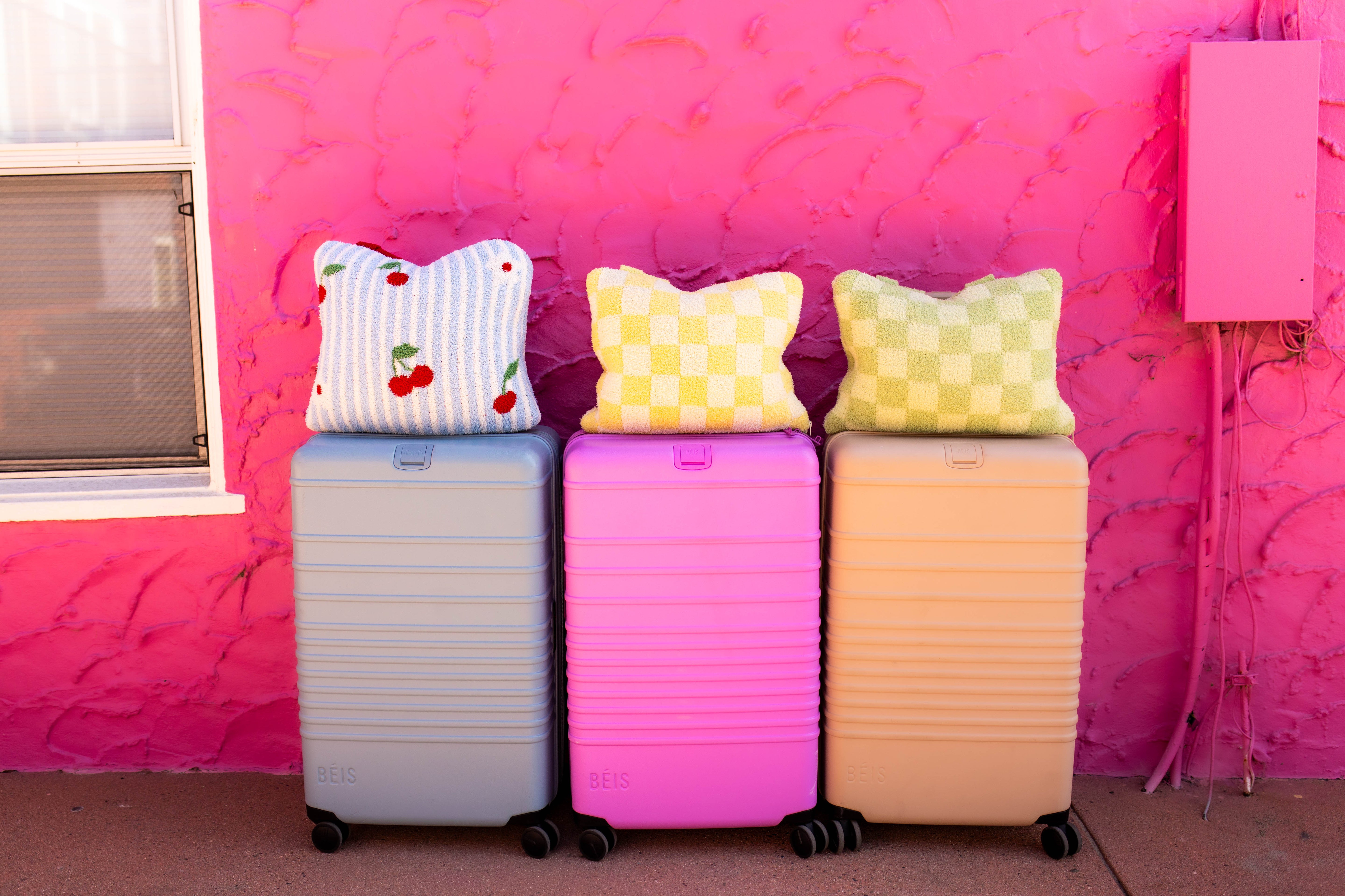 Three hard-shell rolling suitcases in pastel blue, pink, and tan are lined up against a bright pink textured wall, each topped with a quillow.