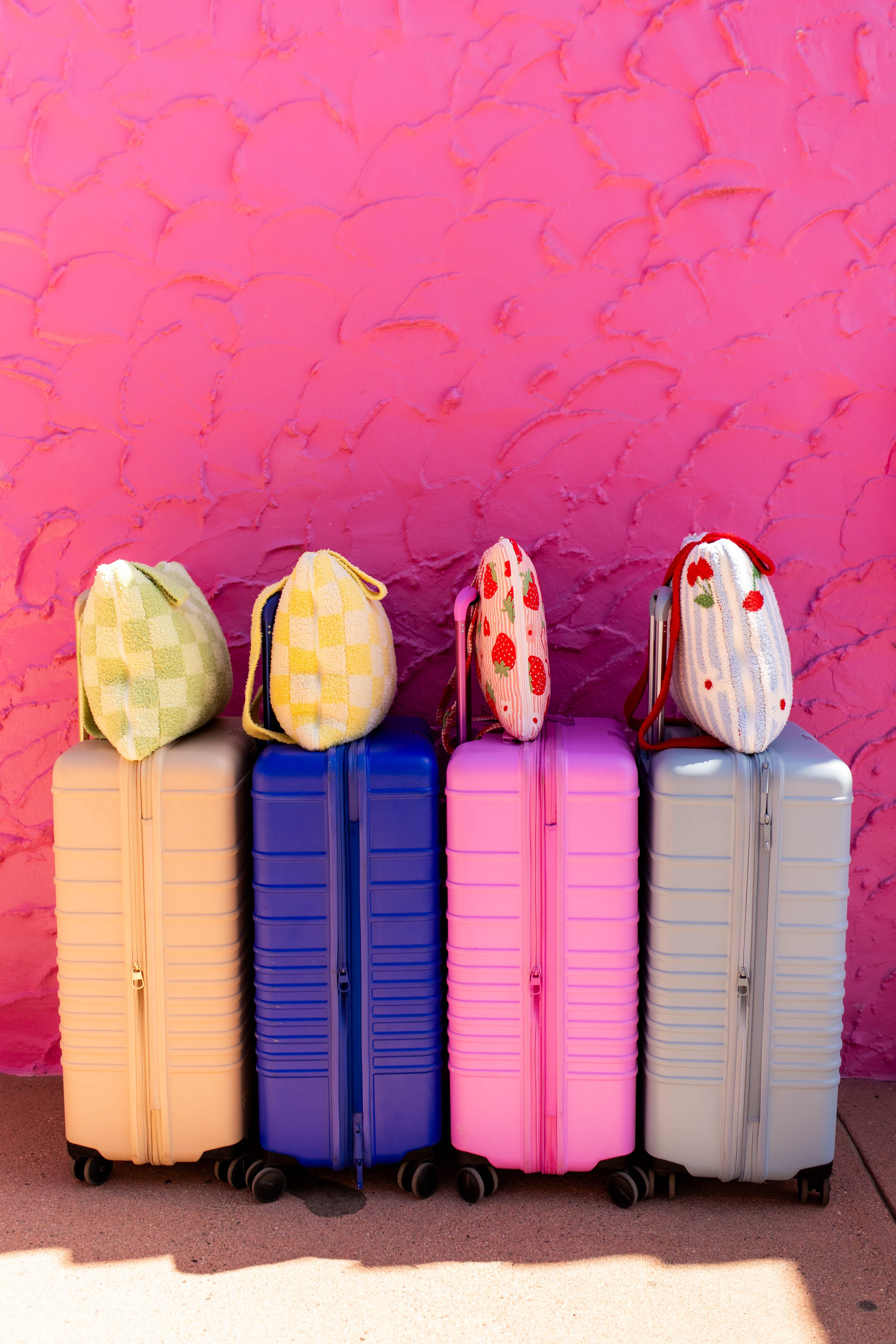 Four hard-shell rolling suitcases in pastel peach, blue, pink, and light blue are lined up against a bright pink textured wall, each topped with a quillow.