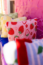 A close-up of a pink striped pillow with a red strawberry pattern, resting on a blue surface in front of a textured pink wall.