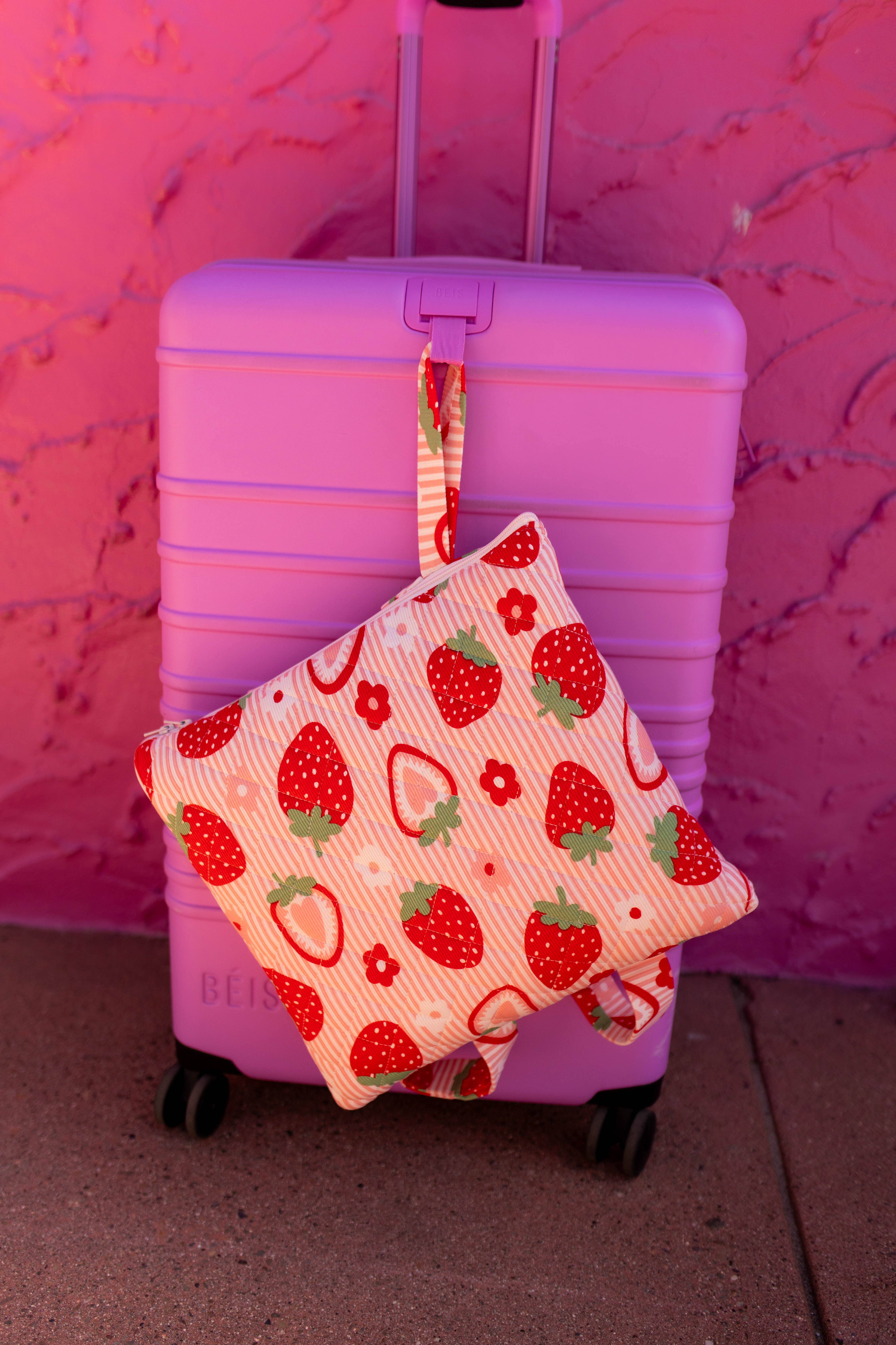 Our outdoor quillow with a strawberry and hanging from a pink suitcase against a pink textured wall.