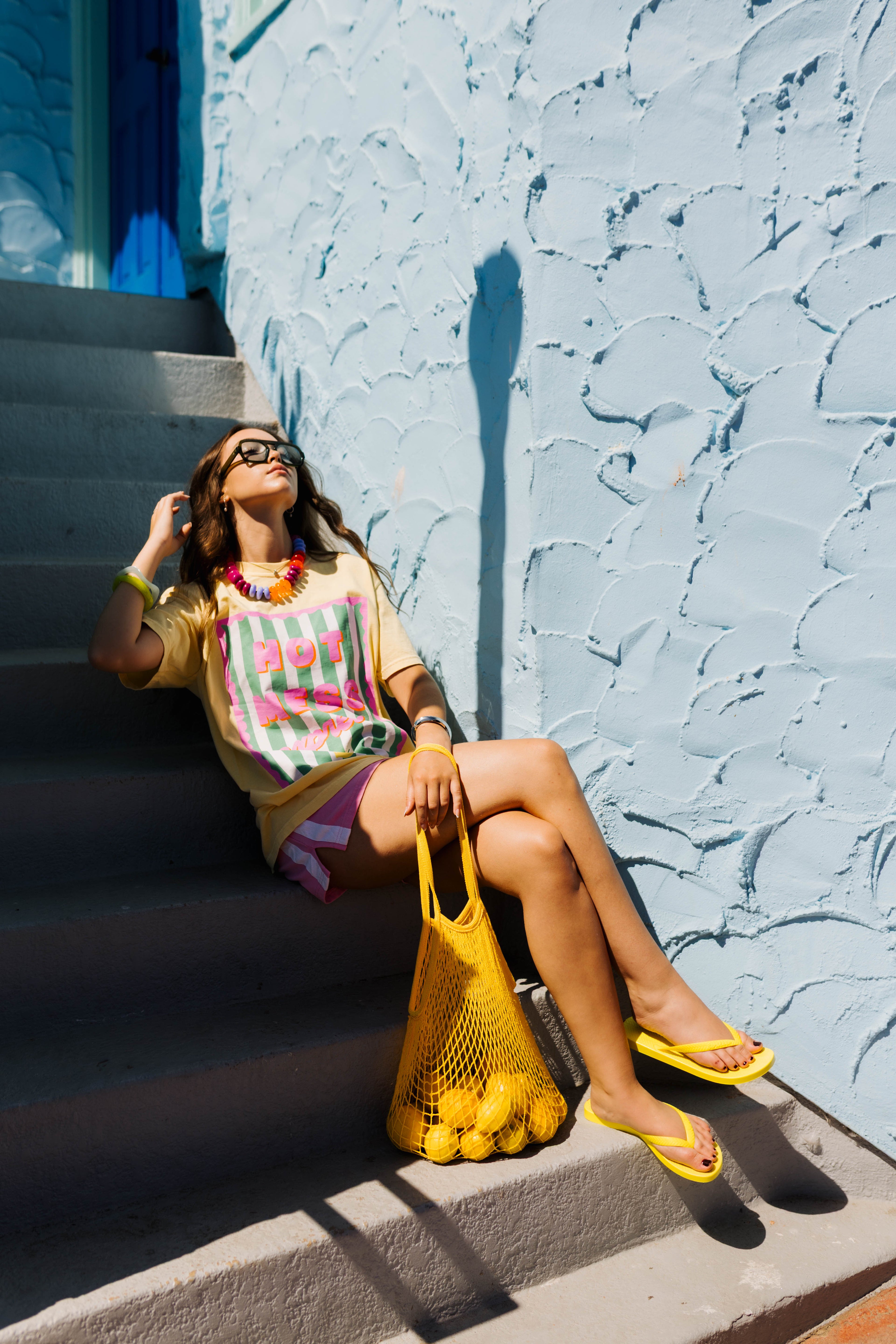 A woman sits on outdoor concrete stairs against a textured light blue wall, wearing a colorful striped graphic T-shirt, pink shorts, yellow flip-flops, and holding a yellow mesh bag of lemons.