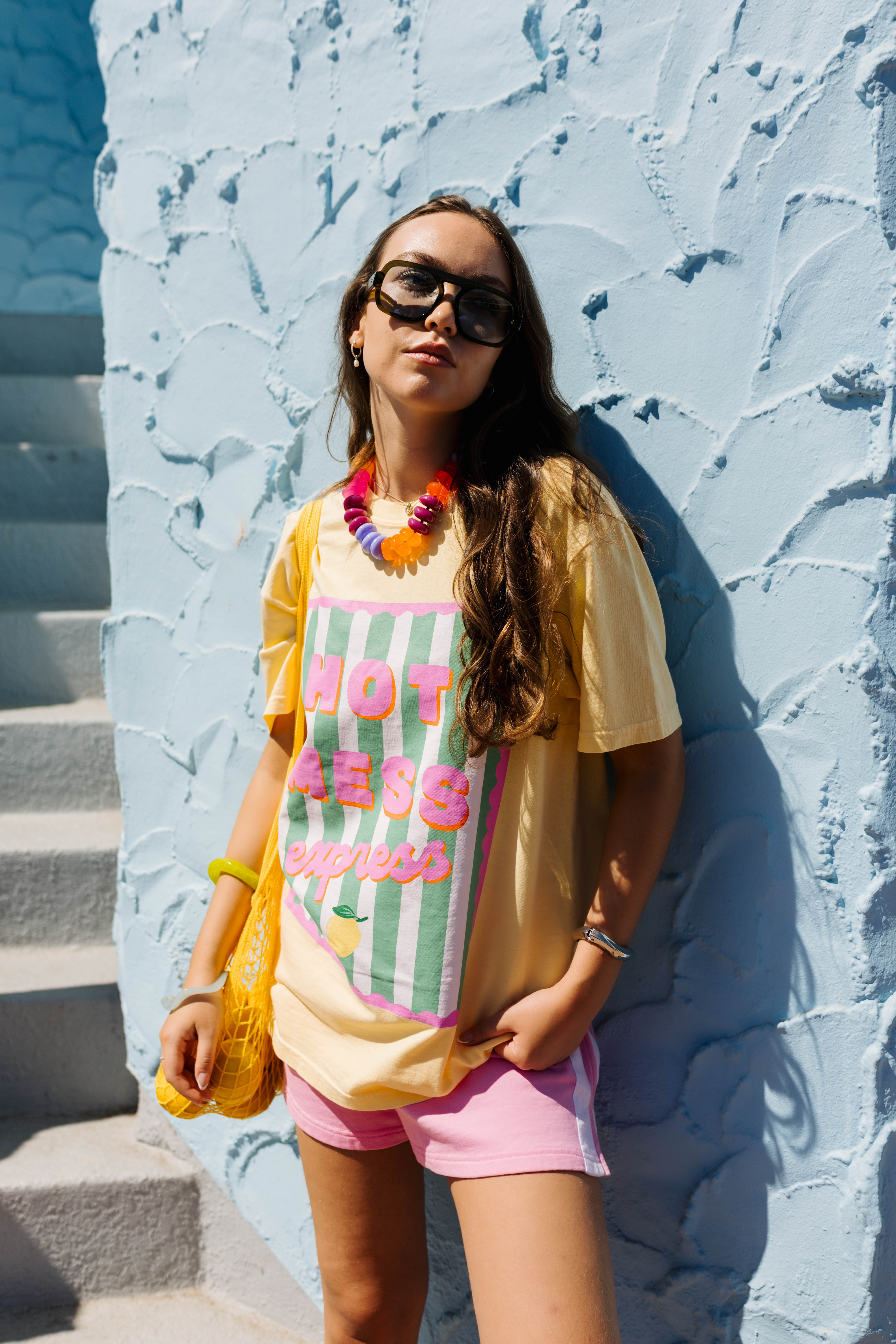 A woman wearing a yellow graphic t-shirt with pink and green text, pink shorts, and a colorful beaded necklace, posing against a textured light blue wall.