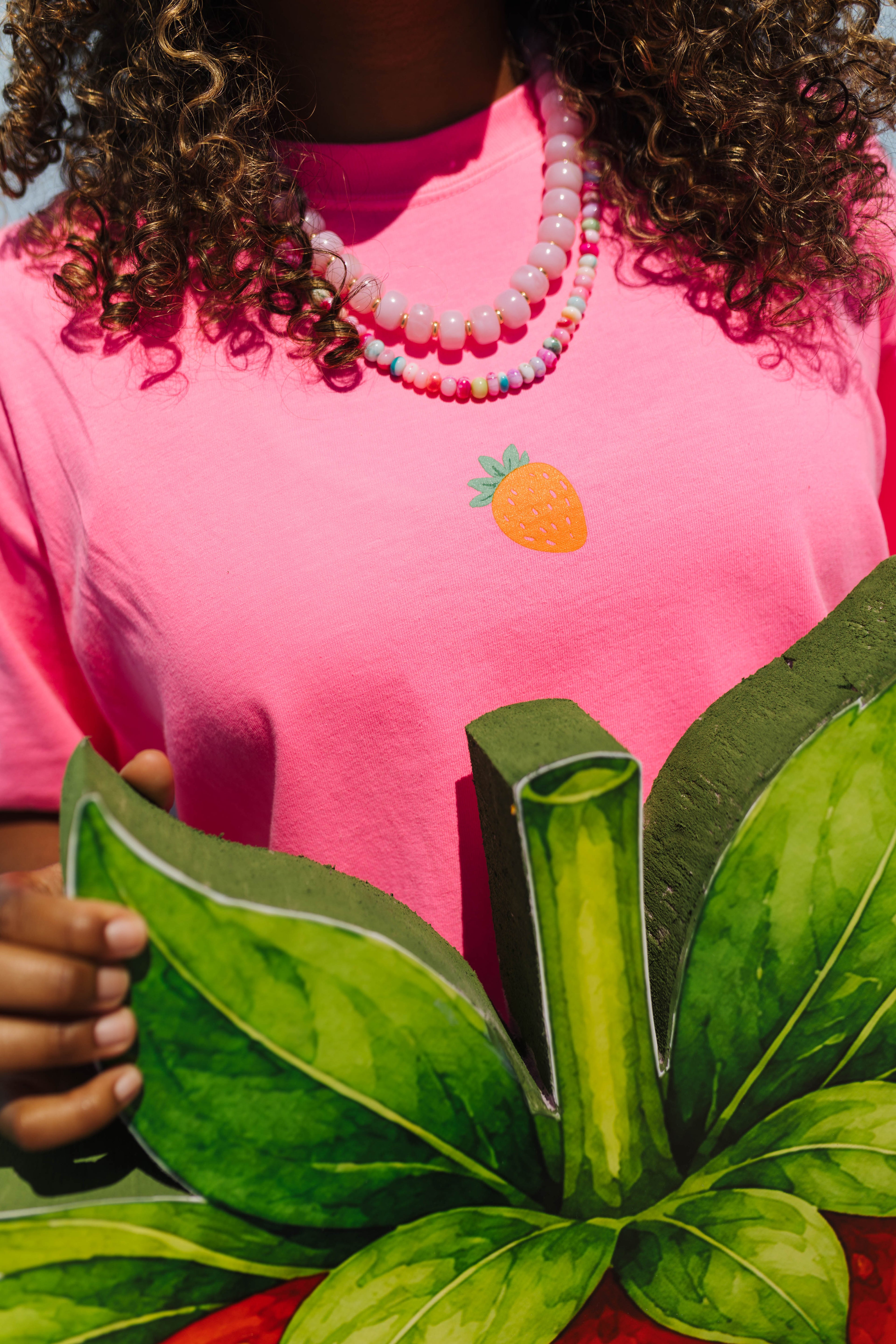 A close-up of a person wearing a bright pink t-shirt with a small embroidered strawberry graphic on the chest. The person is also wearing a layered beaded necklace and holding a large green leafy cutout prop.