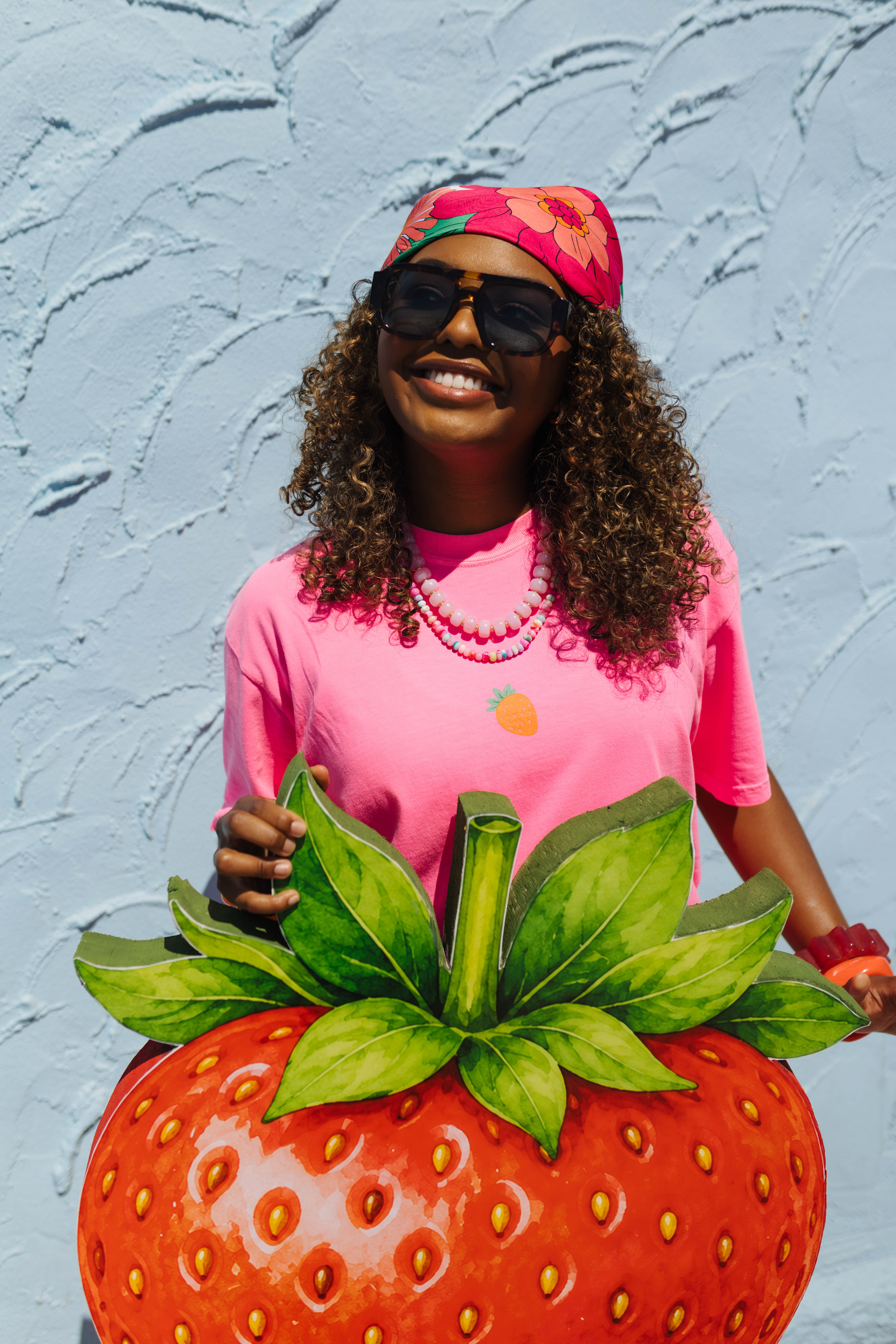 A woman wearing a pink t-shirt with a pineapple graphic, a pink floral headband, and sunglasses, holding a giant strawberry sculpture against a light blue textured wall.