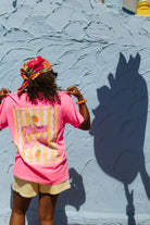 A person wearing a bright pink graphic t-shirt and a colorful headwrap stands with their back to the camera, posing in front of a textured light blue wall that casts a large shadow of the strawberry