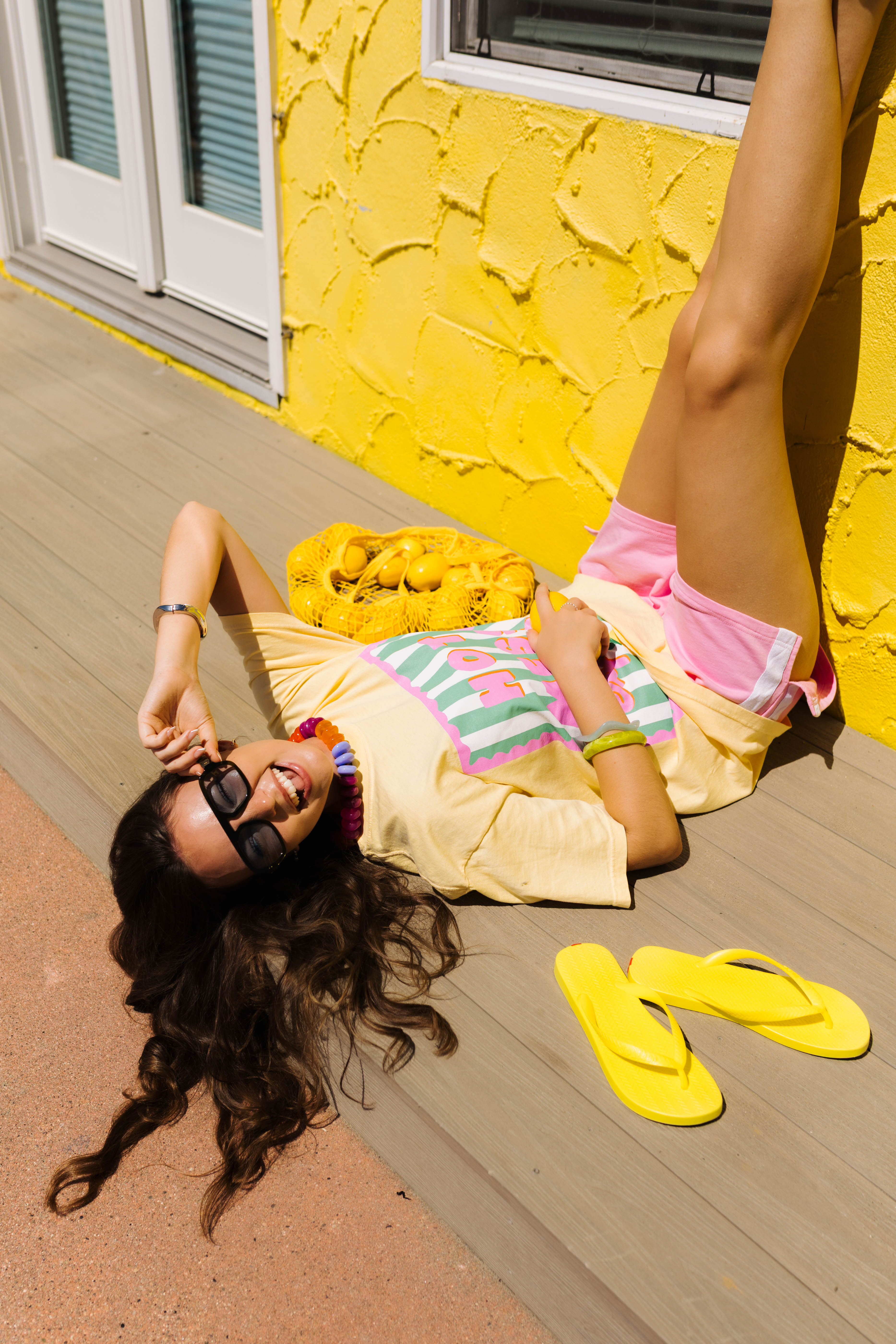 A woman lies on a wooden deck wearing a yellow t-shirt, a striped tank top, pink shorts, and yellow flip-flops, with a matching yellow handbag and a bright yellow stucco wall in the background.
