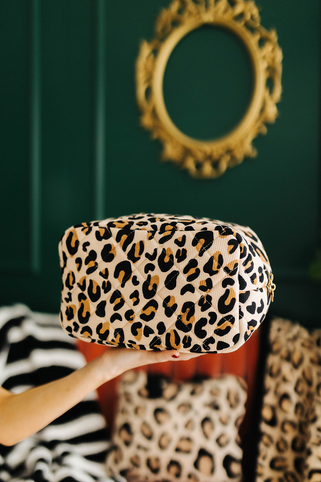 Leopard print cosmetic bag held by a hand against a green wall with a decorative mirror.