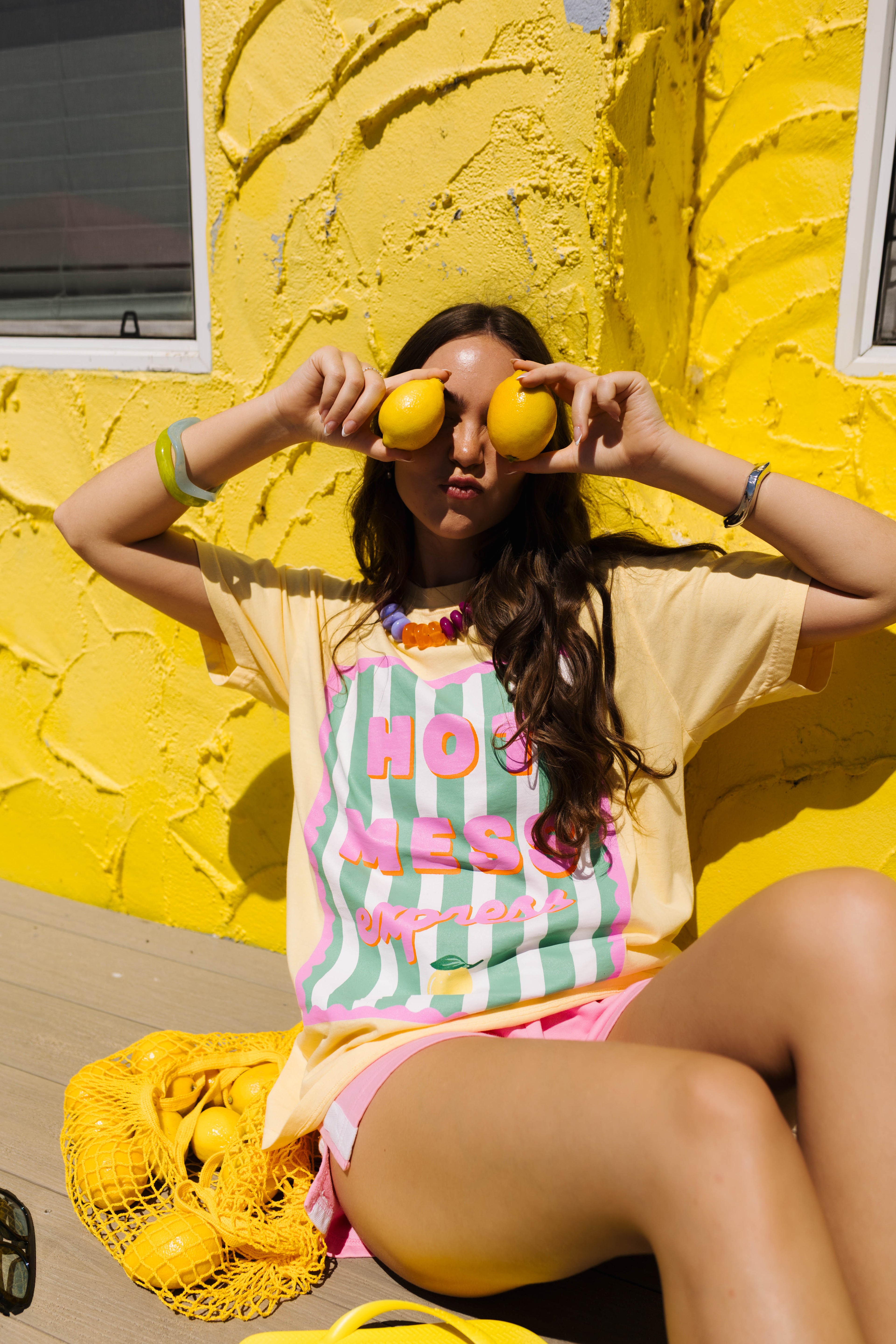 A woman sitting against a bright yellow stucco wall, wearing a pale yellow graphic T-shirt with pink and mint green stripes and the words 'HOT MESS' in pink script. She is holding two yellow lemons in front of her eyes, with a yellow mesh bag of lemons on the floor beside her.