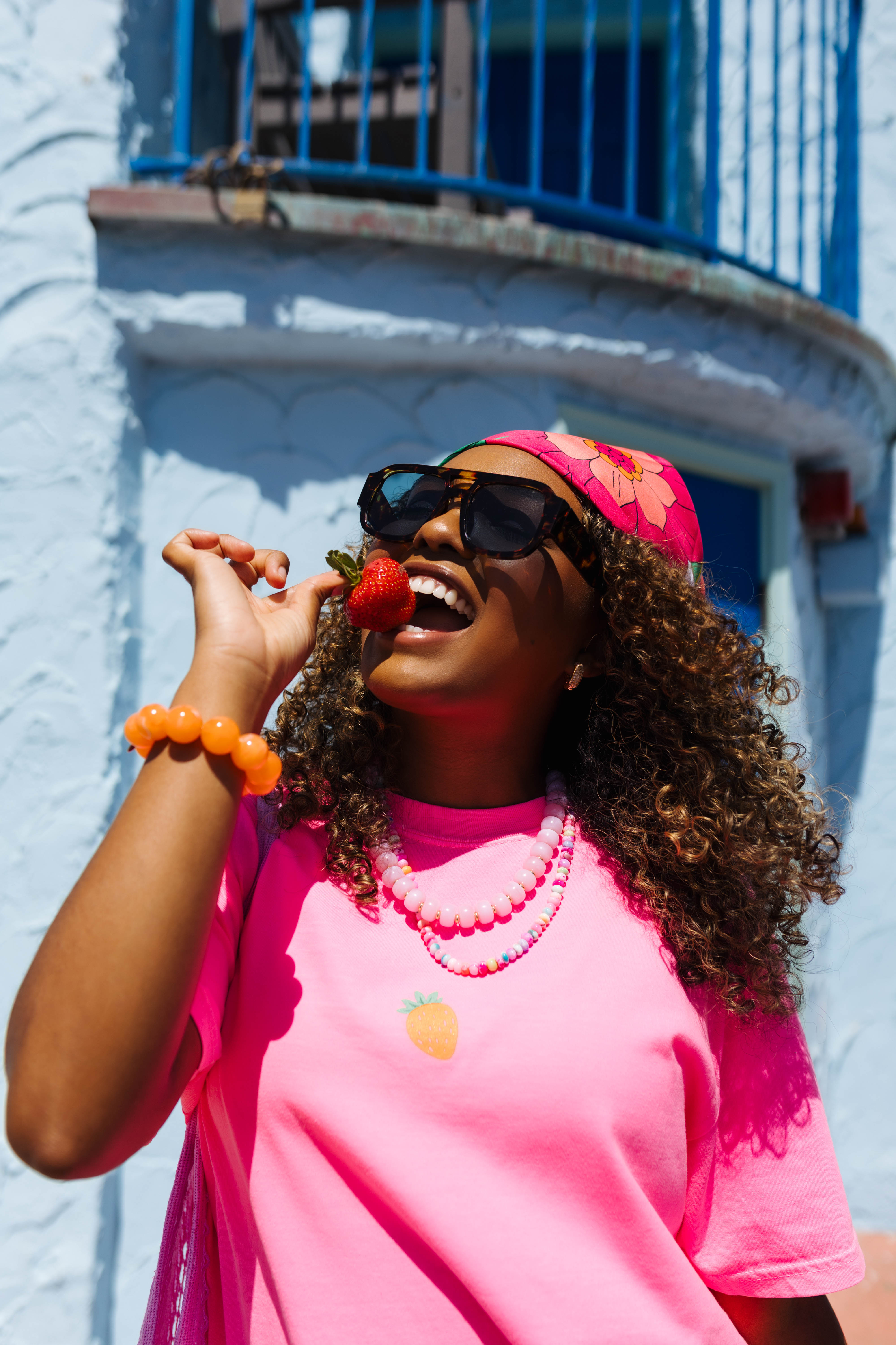 A woman wearing a bright pink t-shirt, matching pink headband, and sunglasses, holding a strawberry near her mouth. She is posed outdoors in front of a light blue stucco building with a blue metal balcony.
