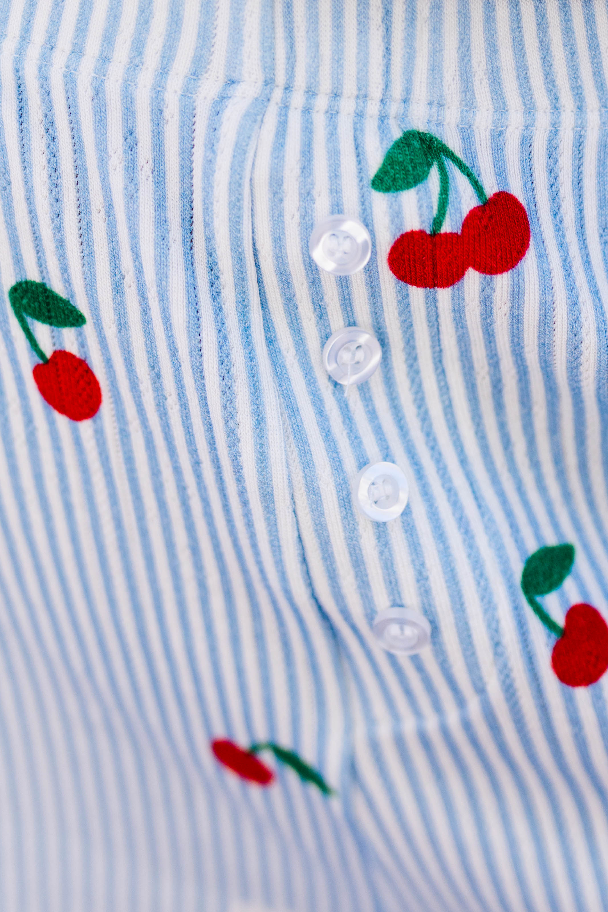 Close-up of a light blue and white striped fabric with printed red cherries and green leaves, featuring a row of white buttons.
