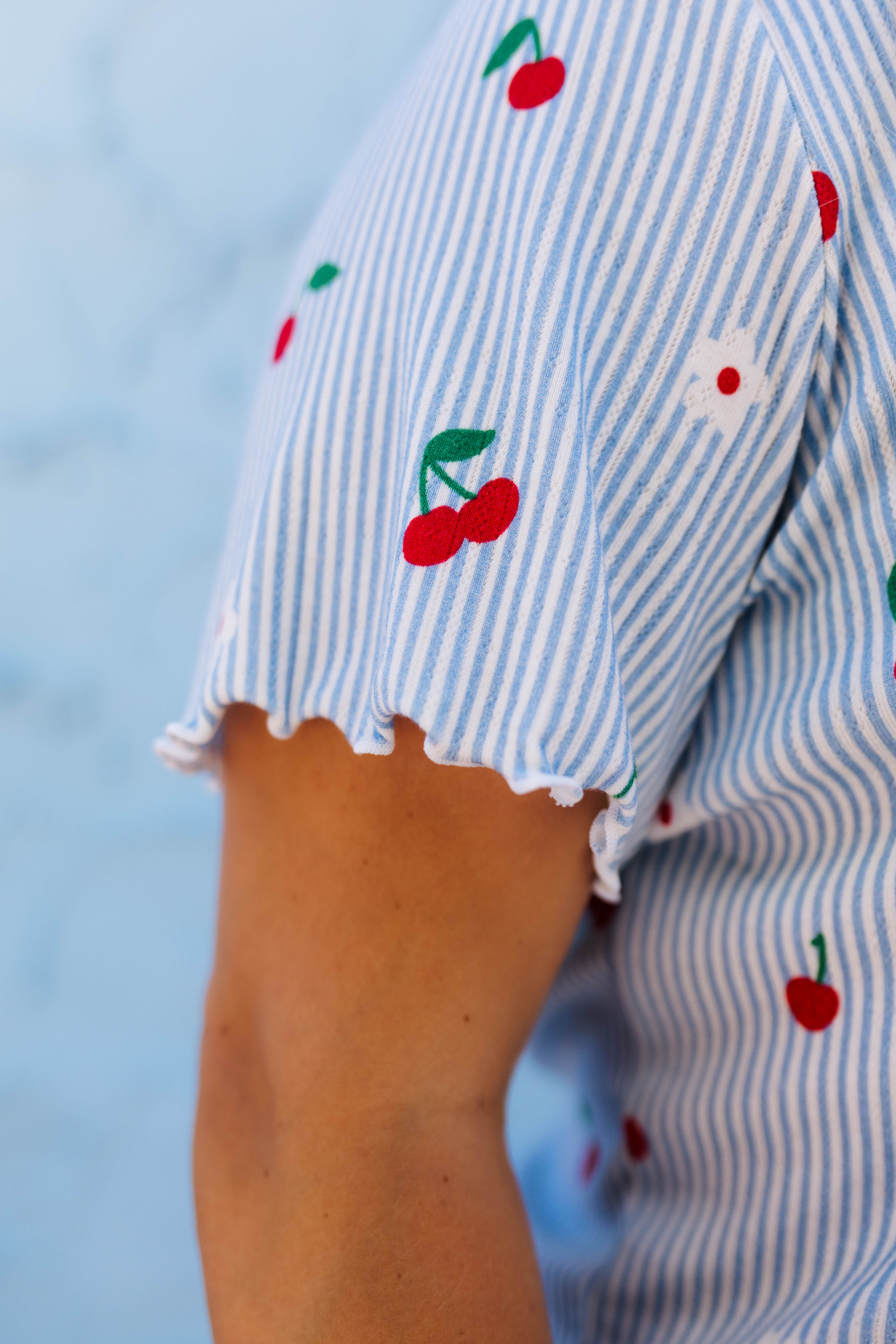 Close-up of a person wearing a light blue and white striped short-sleeve top with printed red cherries and a small white flower on the sleeve.