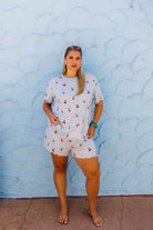 A woman stands barefoot against a light blue textured wall, wearing a matching short-sleeve top and shorts set in white with a cherry print.