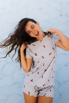 A woman modeling a light blue and white striped short-sleeve pajama set with a cherry and daisy print, posing against a light blue textured wall.