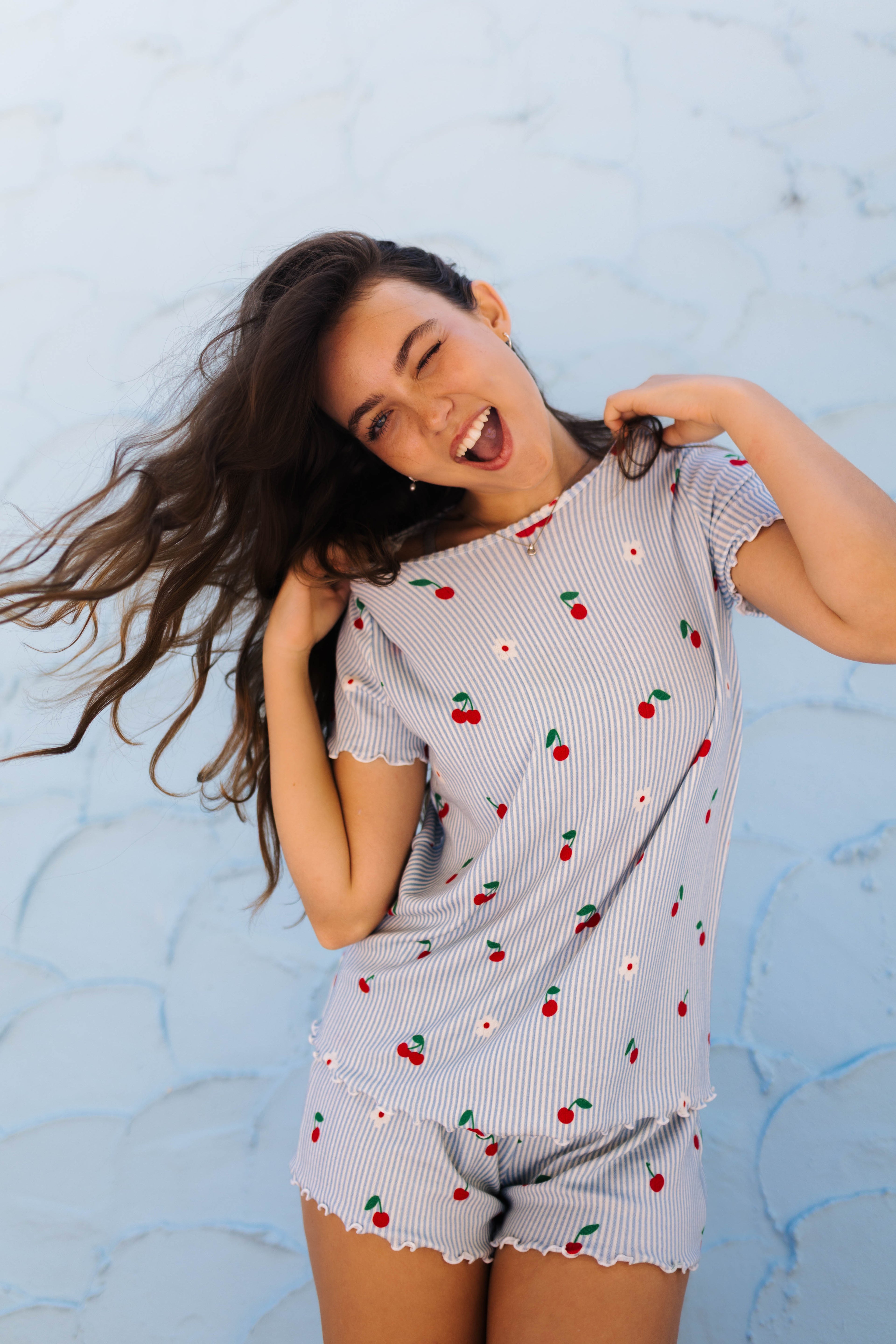 A woman modeling a light blue and white striped short-sleeve pajama set with a cherry and daisy print, posing against a light blue textured wall.