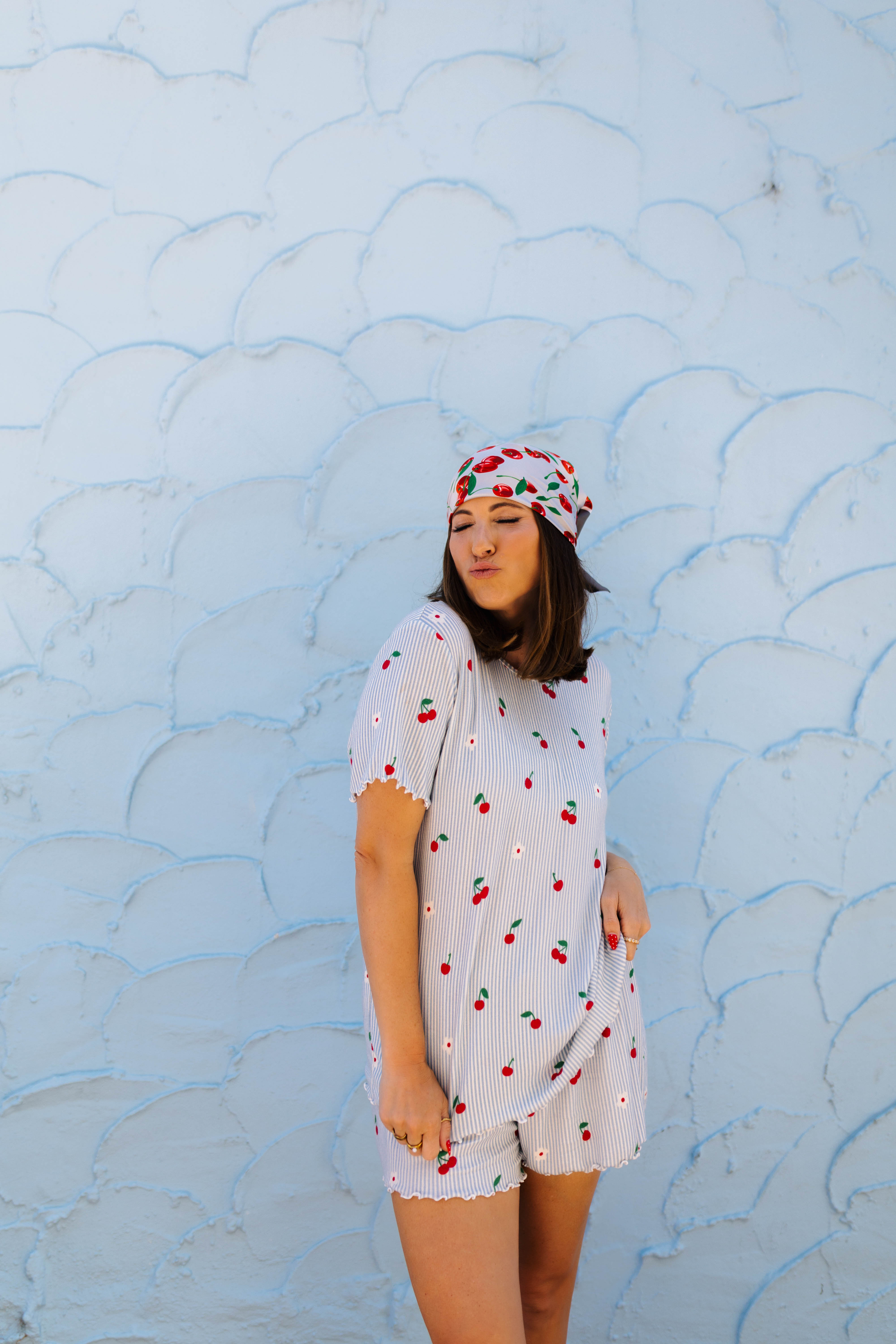 A woman wearing a white cherry-patterned short-sleeve pj and matching headscarf stands against a light blue textured wall.