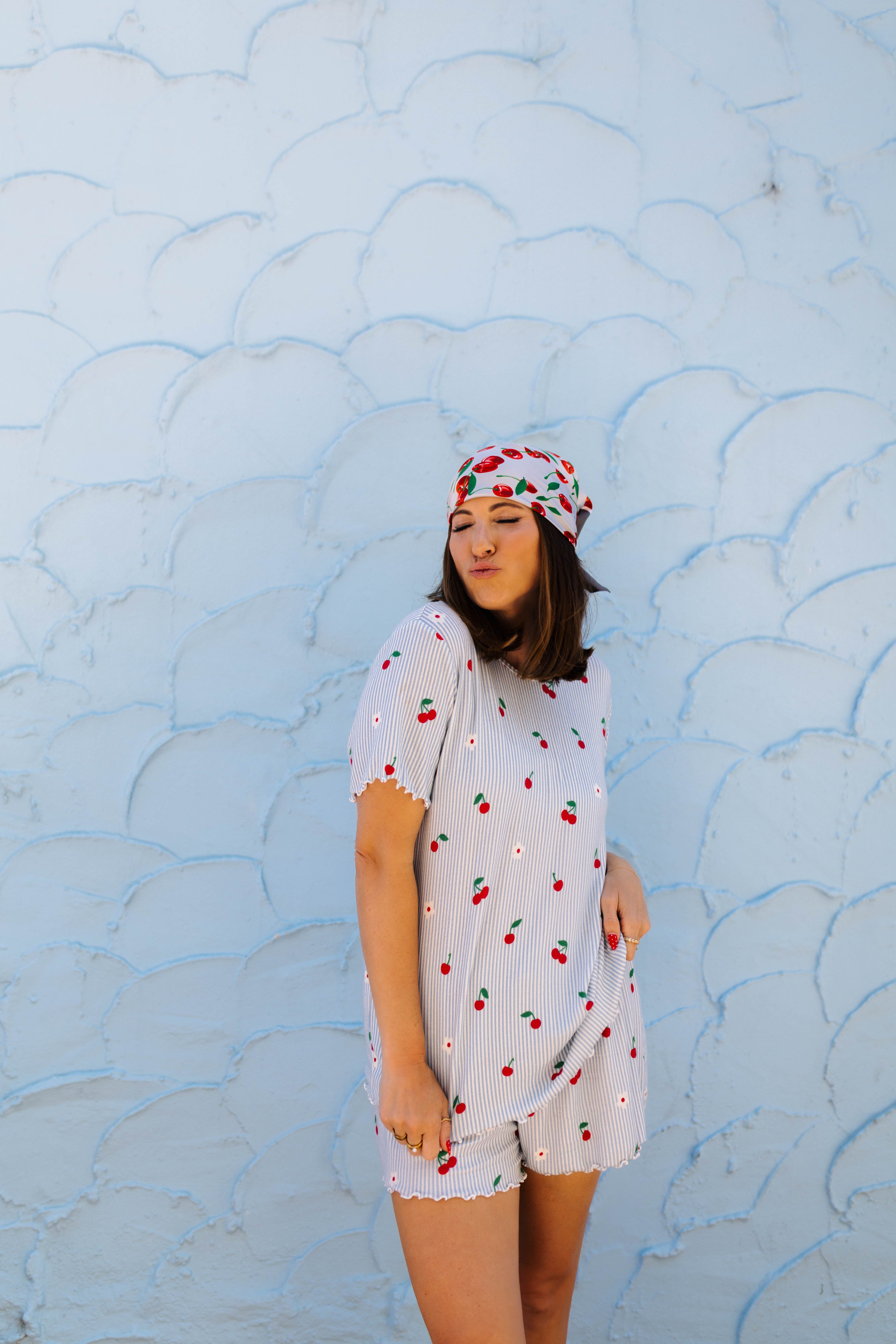 A woman wearing a white cherry-patterned short-sleeve pj and matching headscarf stands against a light blue textured wall.