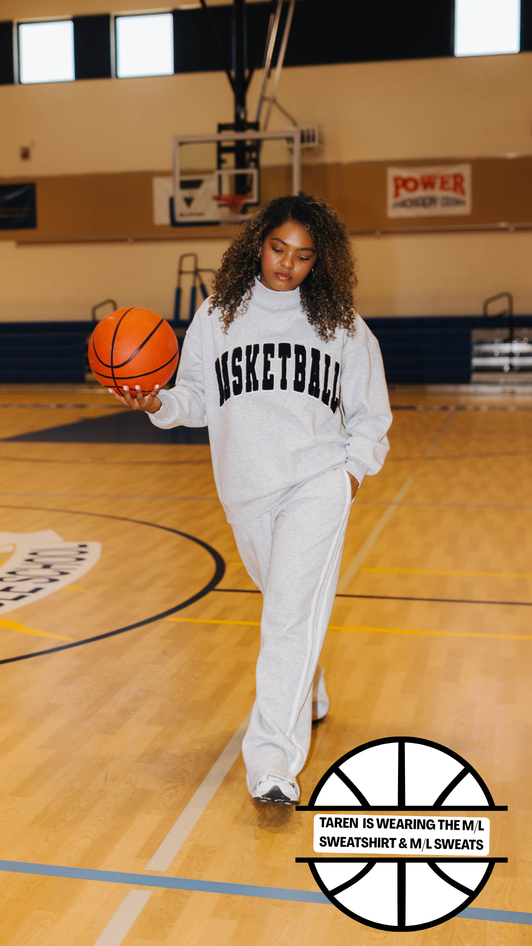 A woman stands on an indoor basketball court wearing a white sweatshirt and sweatpants set with black lettering that reads 'BASKETBALL'. She is holding an orange basketball.