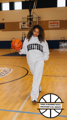 A woman stands on an indoor basketball court wearing a white sweatshirt and sweatpants set with black lettering that reads 'BASKETBALL'. She is holding an orange basketball.