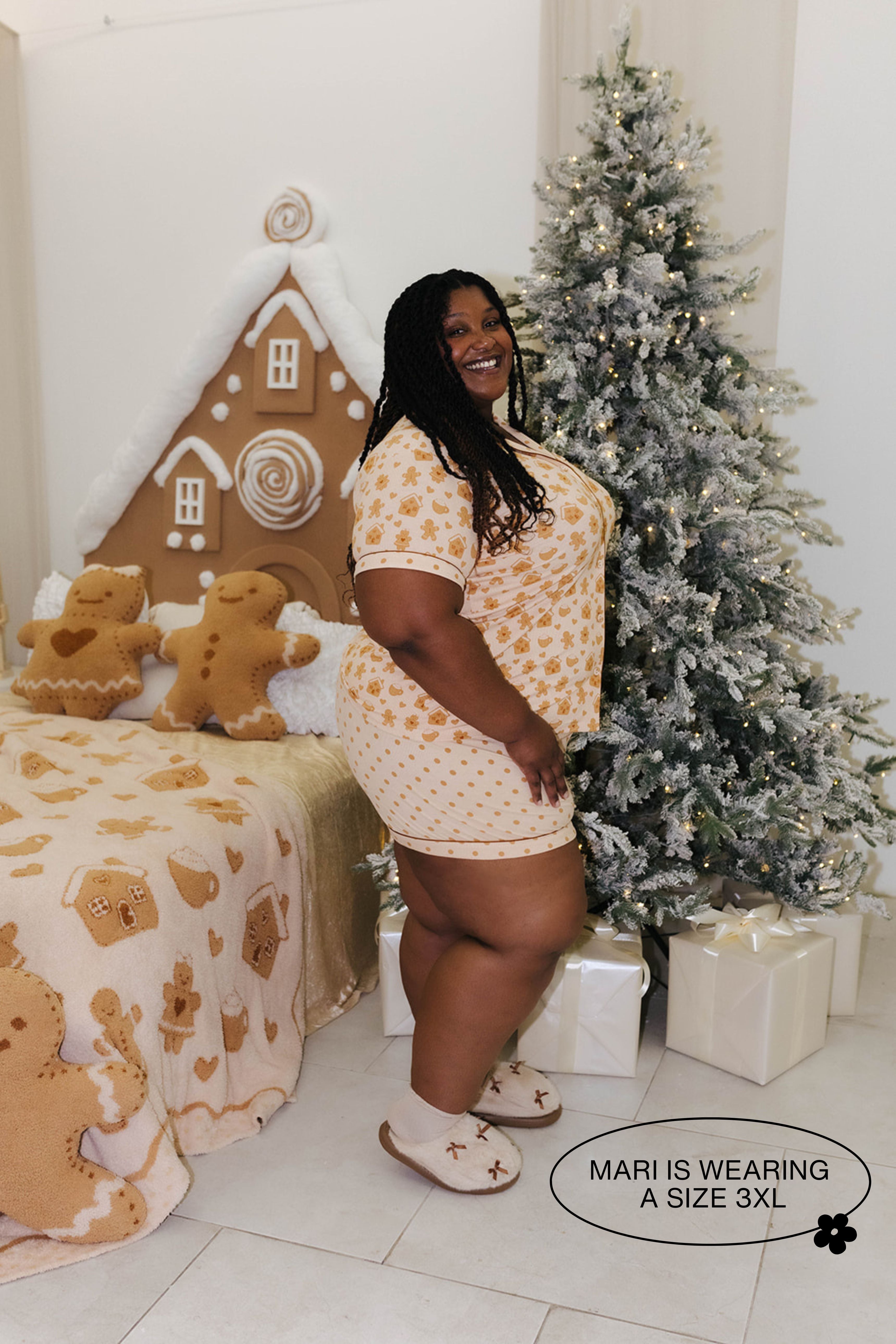 Woman in a pajama set standing next to a decorated Christmas tree and gingerbread-themed bedspread.