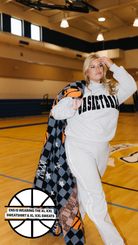 A woman posing in a gym on a basketball court wearing a gray sweatshirt and sweatpants set, with a black, gray, and orange checkered blanket draped over her shoulder. The sweatshirt features the word 'BASKETBALL' printed across the chest in black.
