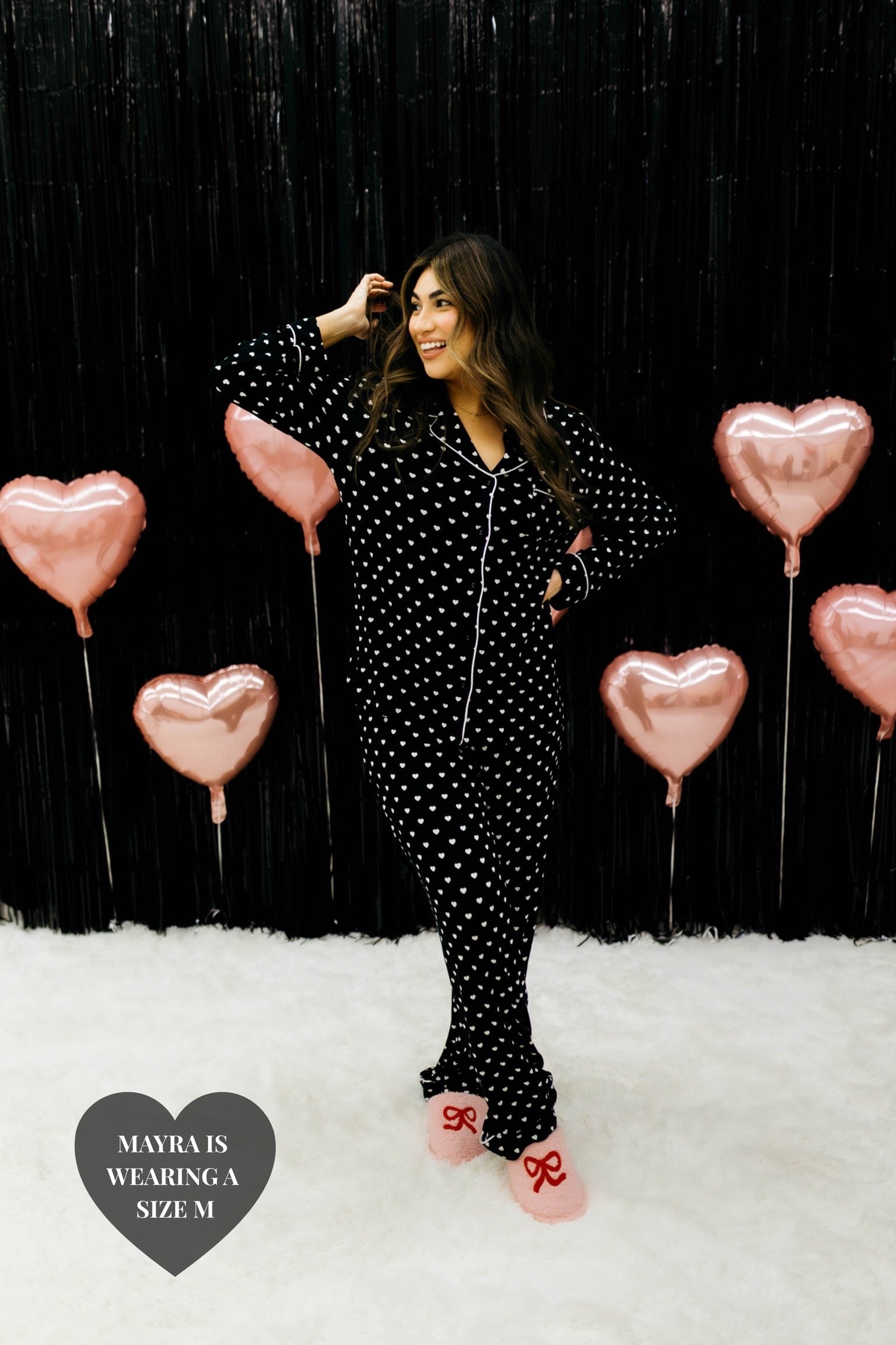 Mayra posing in front of a black wall with pink heart balloons showing the black heart pjs