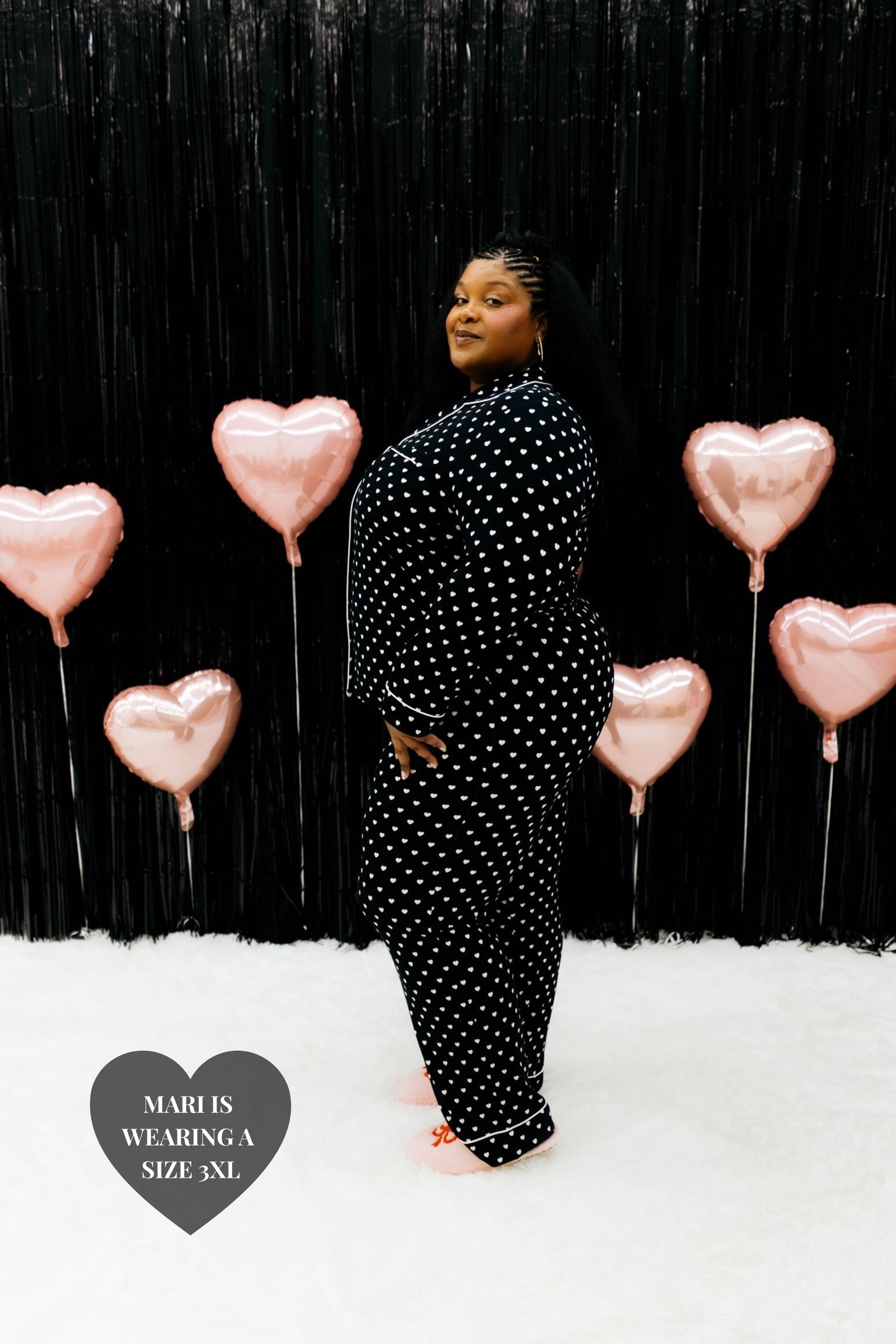 Mari wearing the size 3XL posing to the side and similing standing in front of the black wall with pink hearts 
