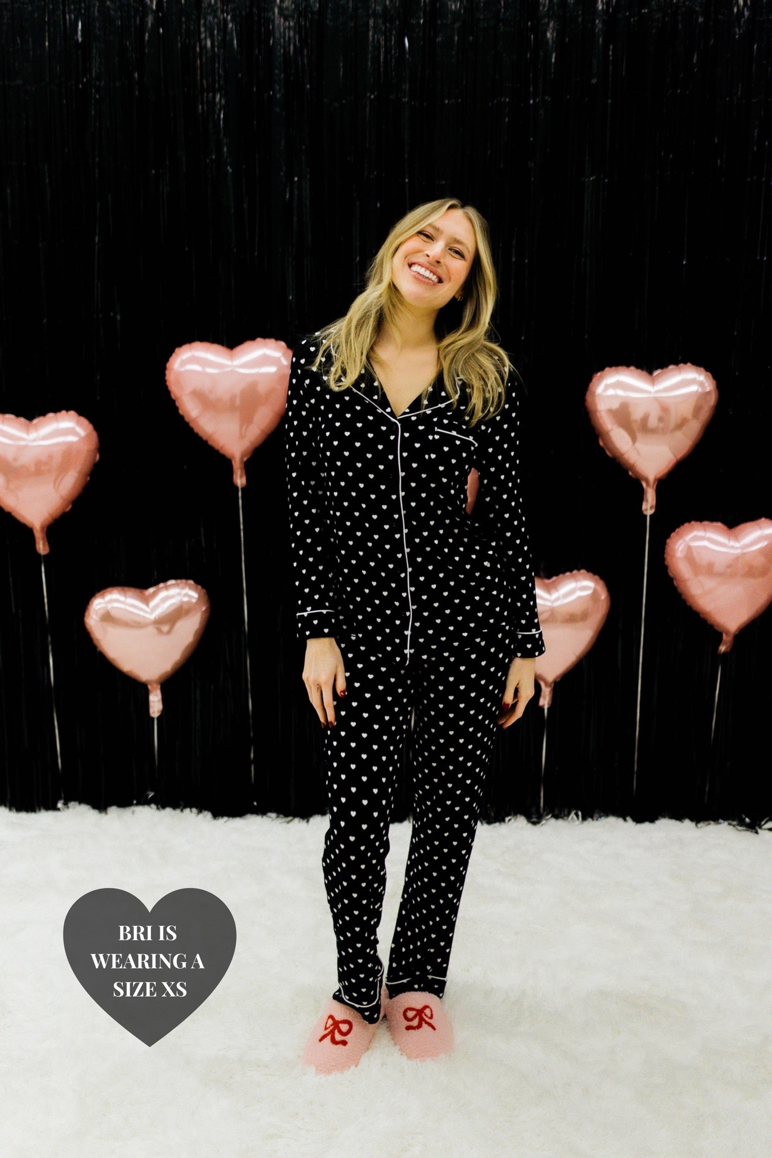 Bri wearing the XS smiling big in front of the black wall showing off the black heart pjs
