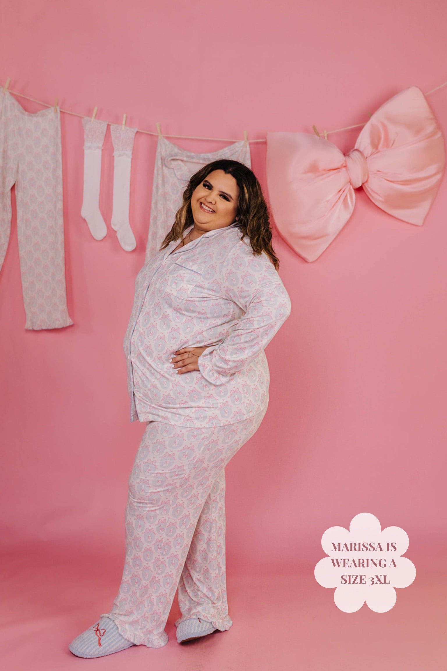 Woman wearing a white pajama set against a pink background with the pajamas hung and a big pink bow. 